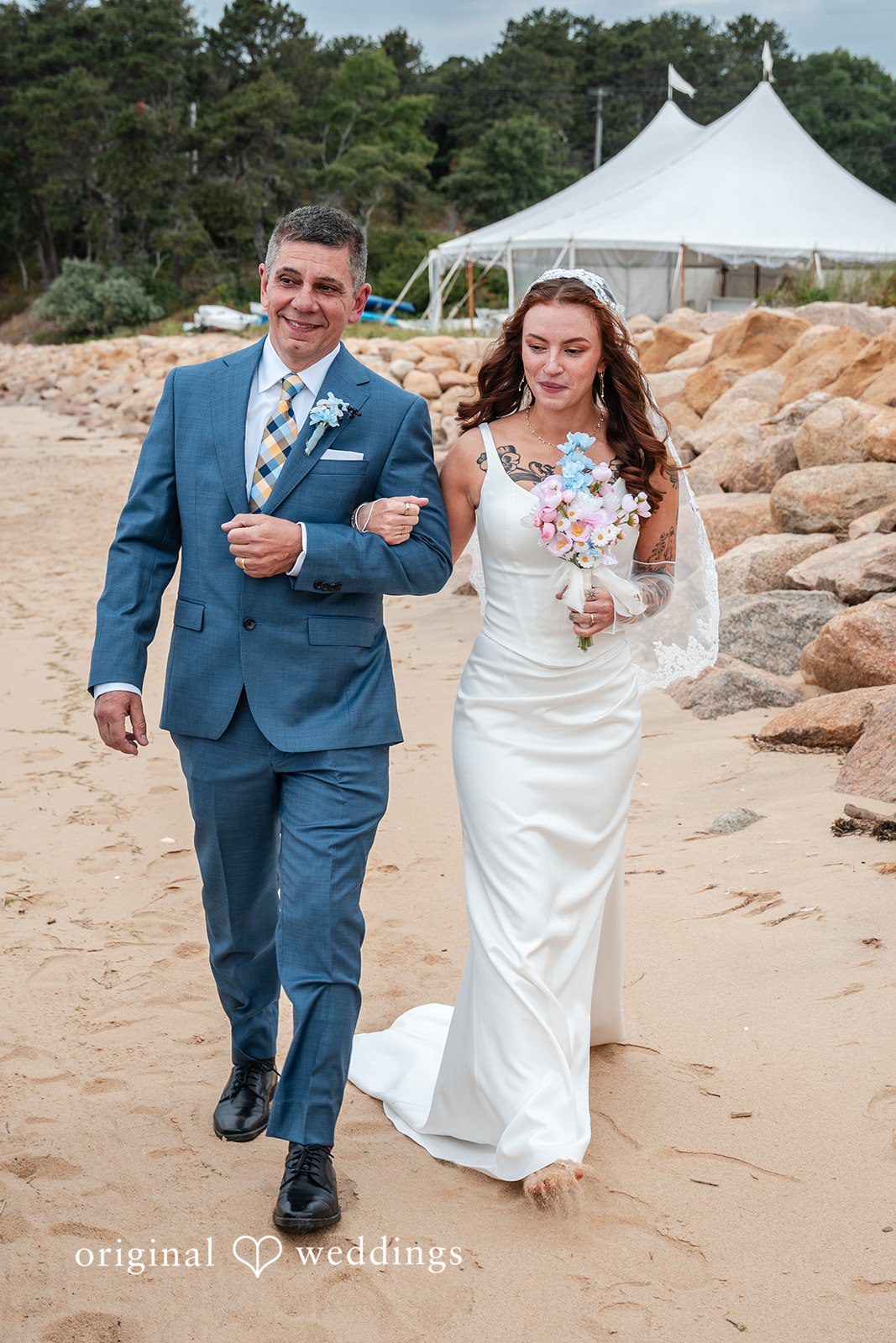 Jacqueline + Sarah A man and bride walking hand-in-hand and smiling down a sandy pathway.