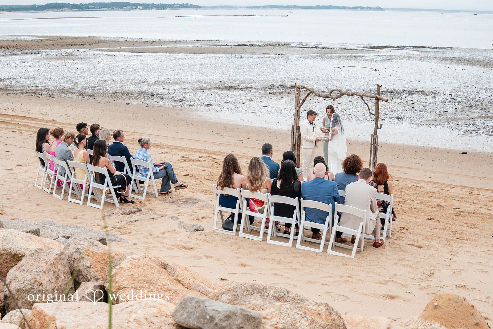 Jacqueline + Sarah An aerial view of a wedding ceremony set up with chairs on a beach.