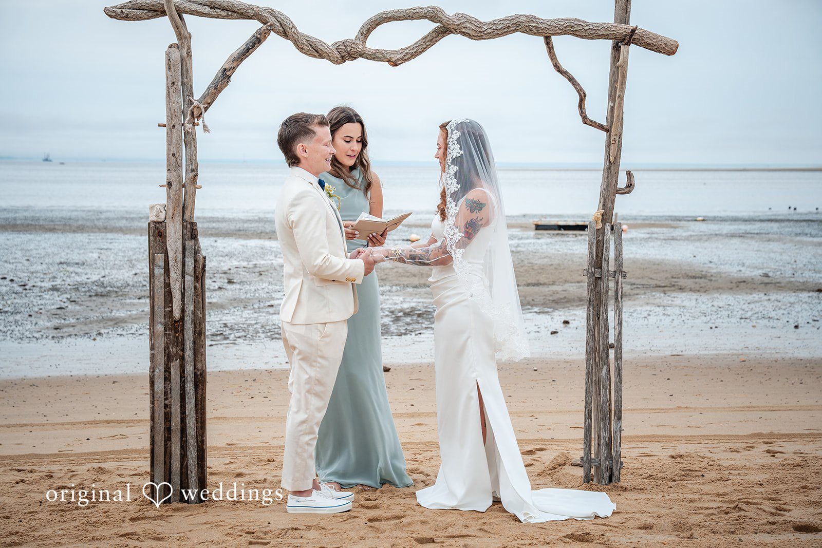 Jacqueline + Sarah A wedding couple standing together under a rustic driftwood arch on the beach.