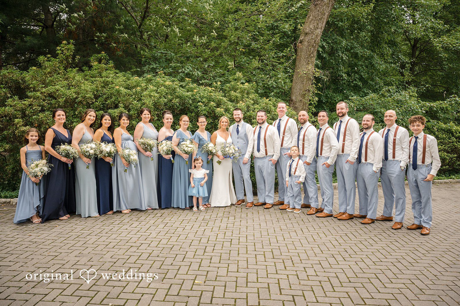 Morgan + Colin A large wedding party in blue attire posing in a wide semicircle on a paved area.