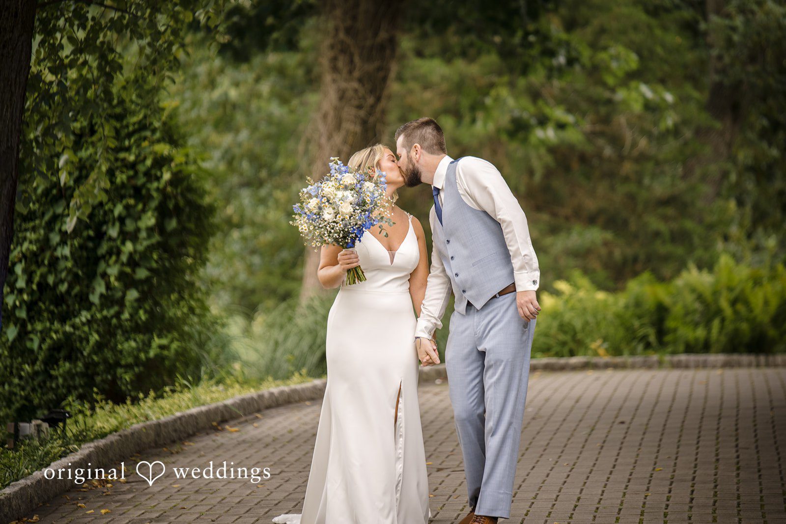 Morgan + Colin A bride and groom kissing lovingly on a path framed by lush trees.