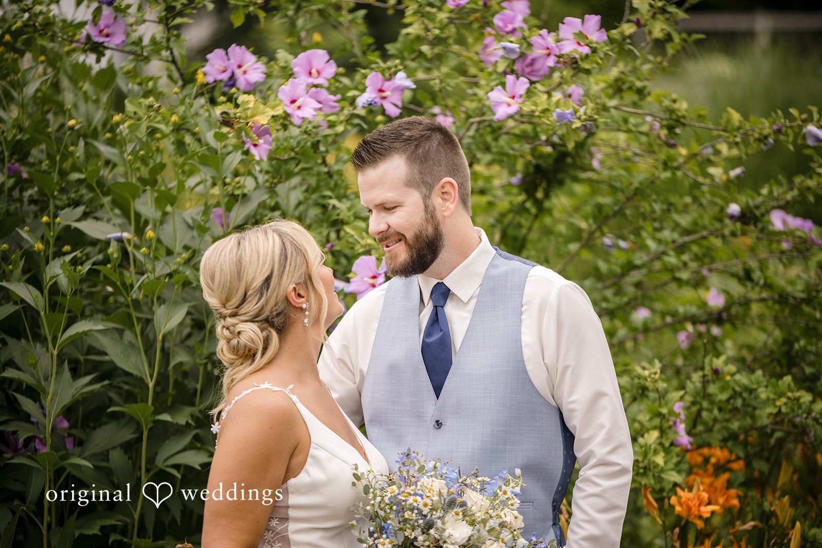 Morgan + Colin A close-up of a groom smiling warmly at his bride amid green foliage.