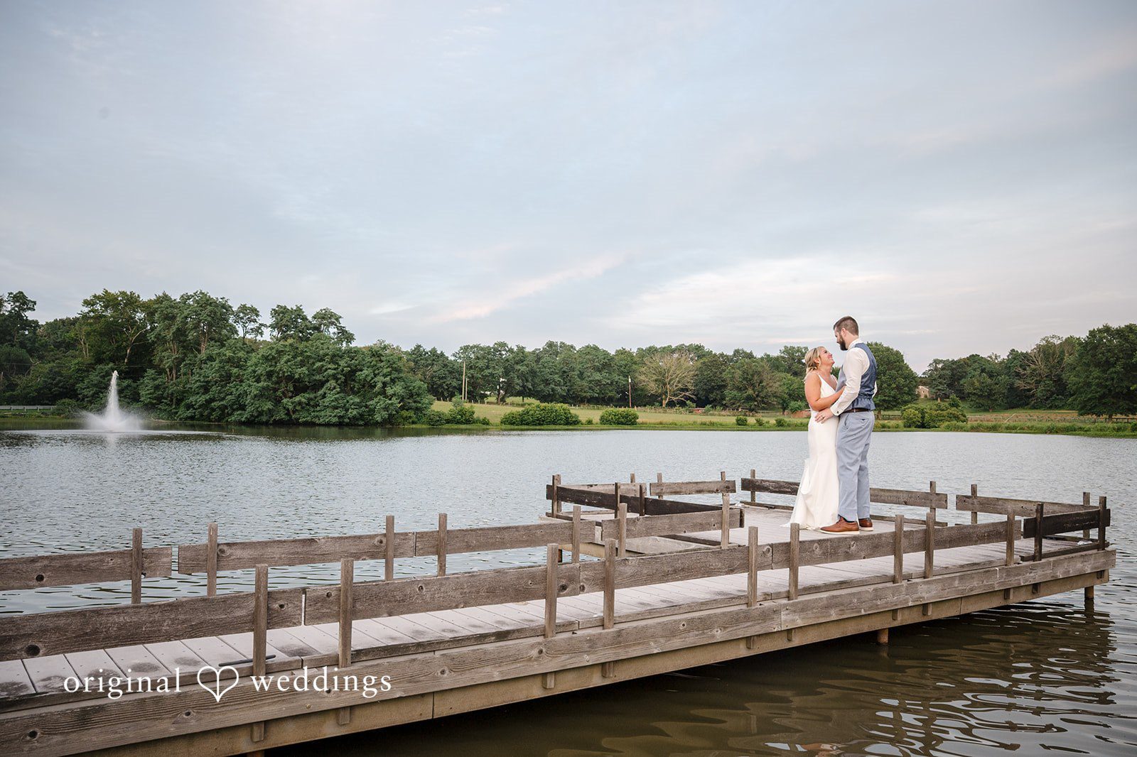 Morgan + Colin A bride and groom standing together at the edge of a wooden dock overlooking a lake.