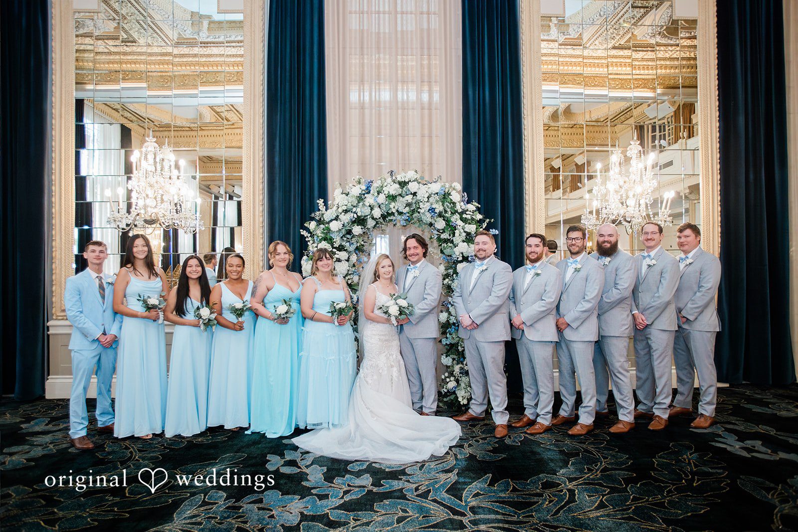 Sara + Michael A large wedding party consisting of bridesmaids in blue dresses and groomsmen in grey suits posing together.