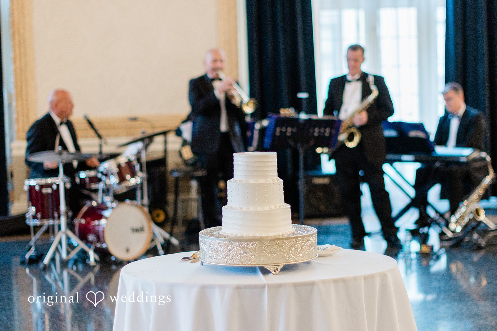 Sara + Michael A multi-tiered white wedding cake displayed on a table while a live band plays behind it.