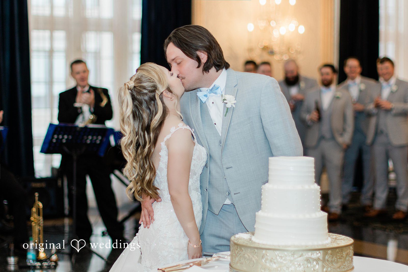 Sara + Michael A groom kissing bride while cutting a white multi-tiered wedding cake at the reception.