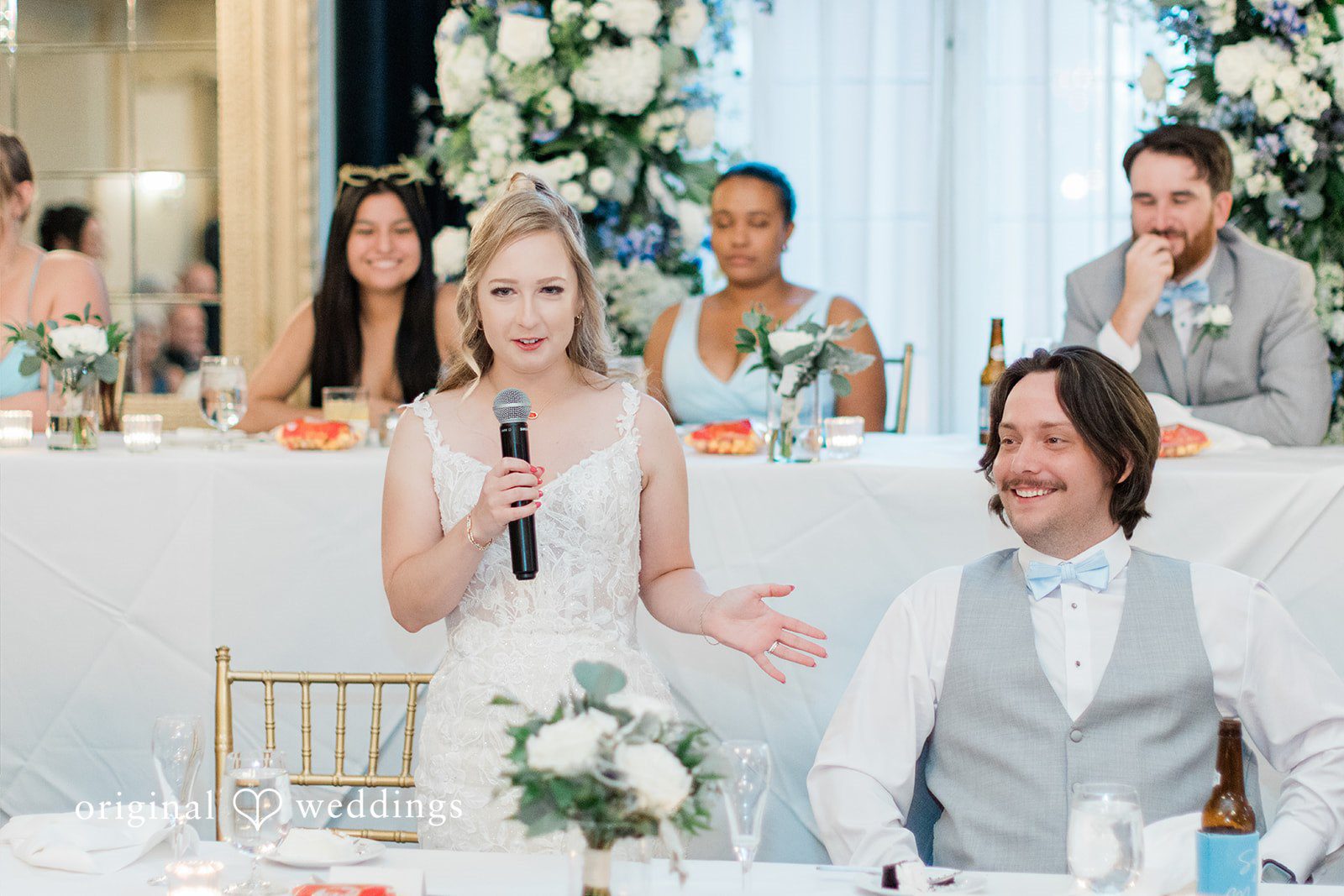 Sara + Michael A bride holding a microphone and speaking at the reception head table while the groom smiles.