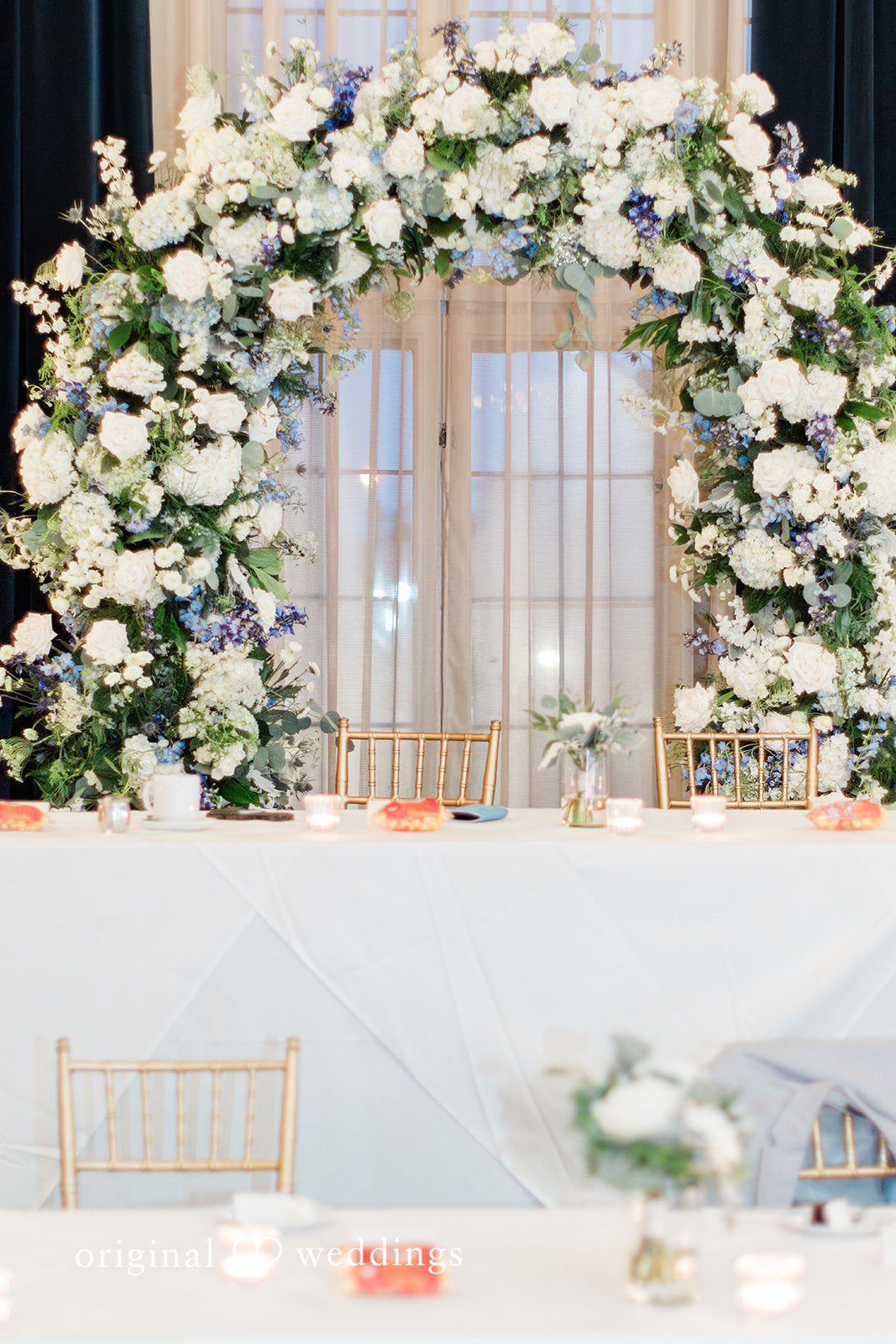 Sara + Michael A long wedding head table decorated with lush white floral arrangements and candles.