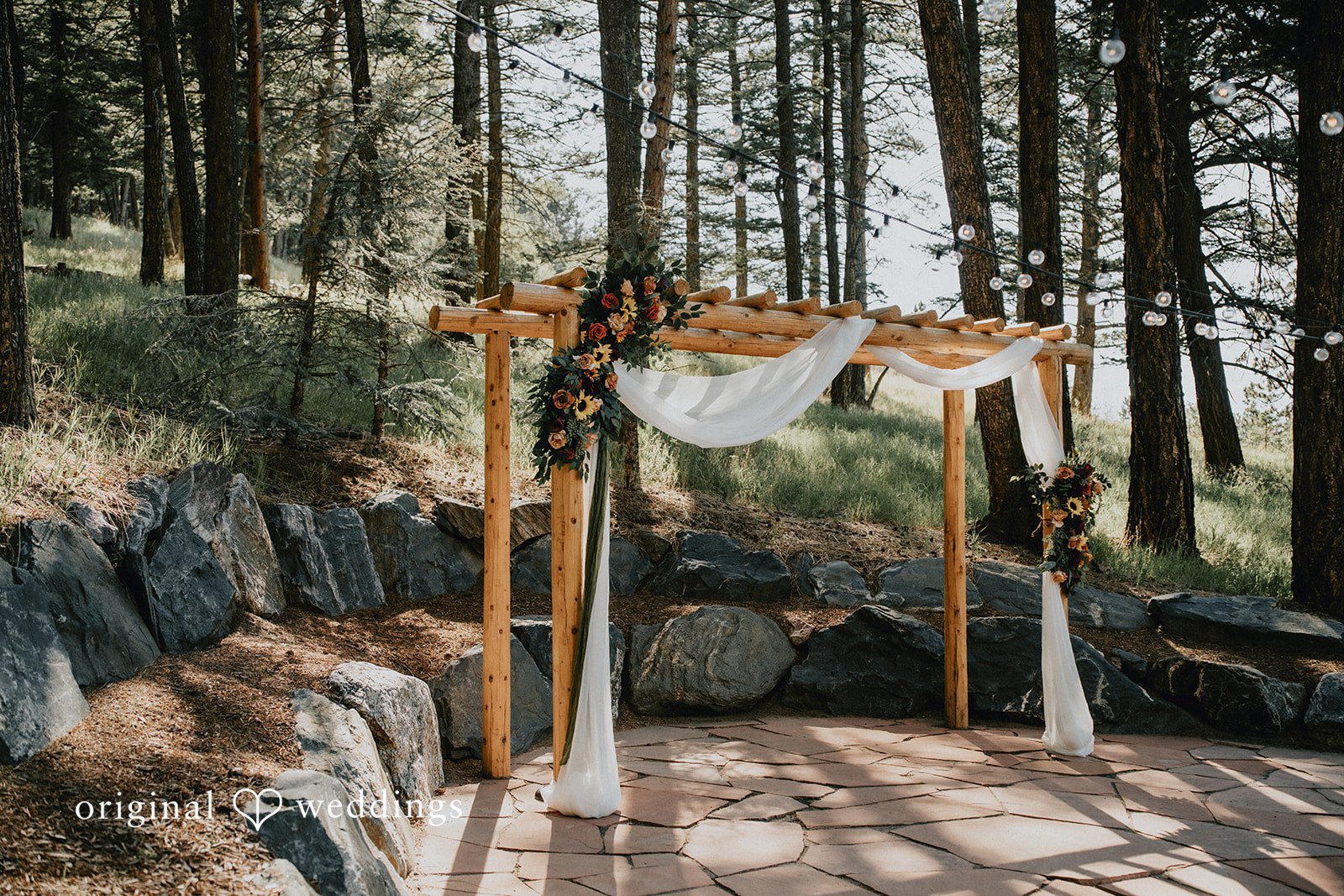 Jennifer + Danielle A wooden wedding arch draped with white fabric stands in a forest setting.