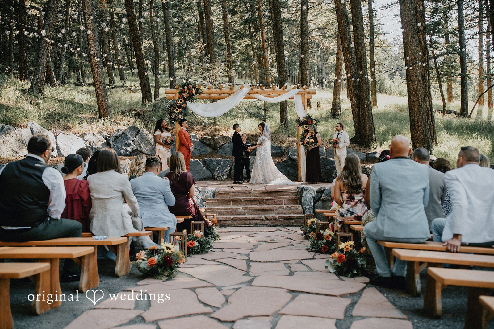 Jennifer + Danielle Rows of seated wedding guests watch a ceremony in a wooded area.