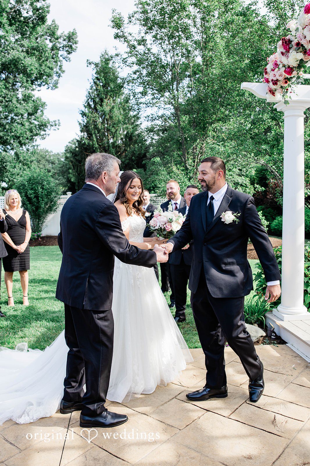 A man in a black suit shakes hands with the groom while giving away the bride at a wedding altar.