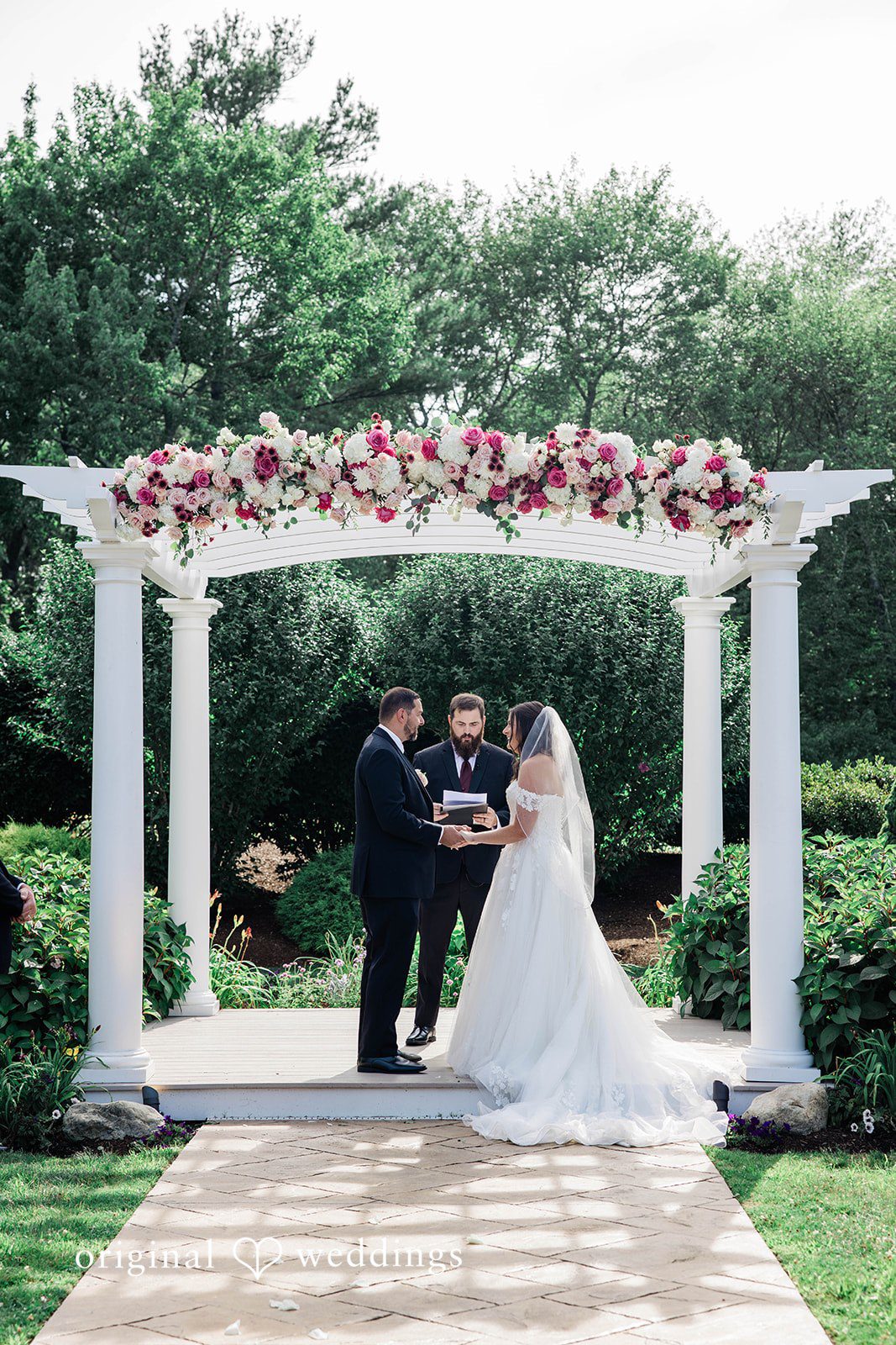 A wedding ceremony taking place beneath a decorated white floral arbor.