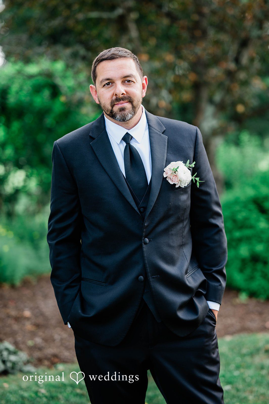 A groom in a black suit posing against a background of green trees.
