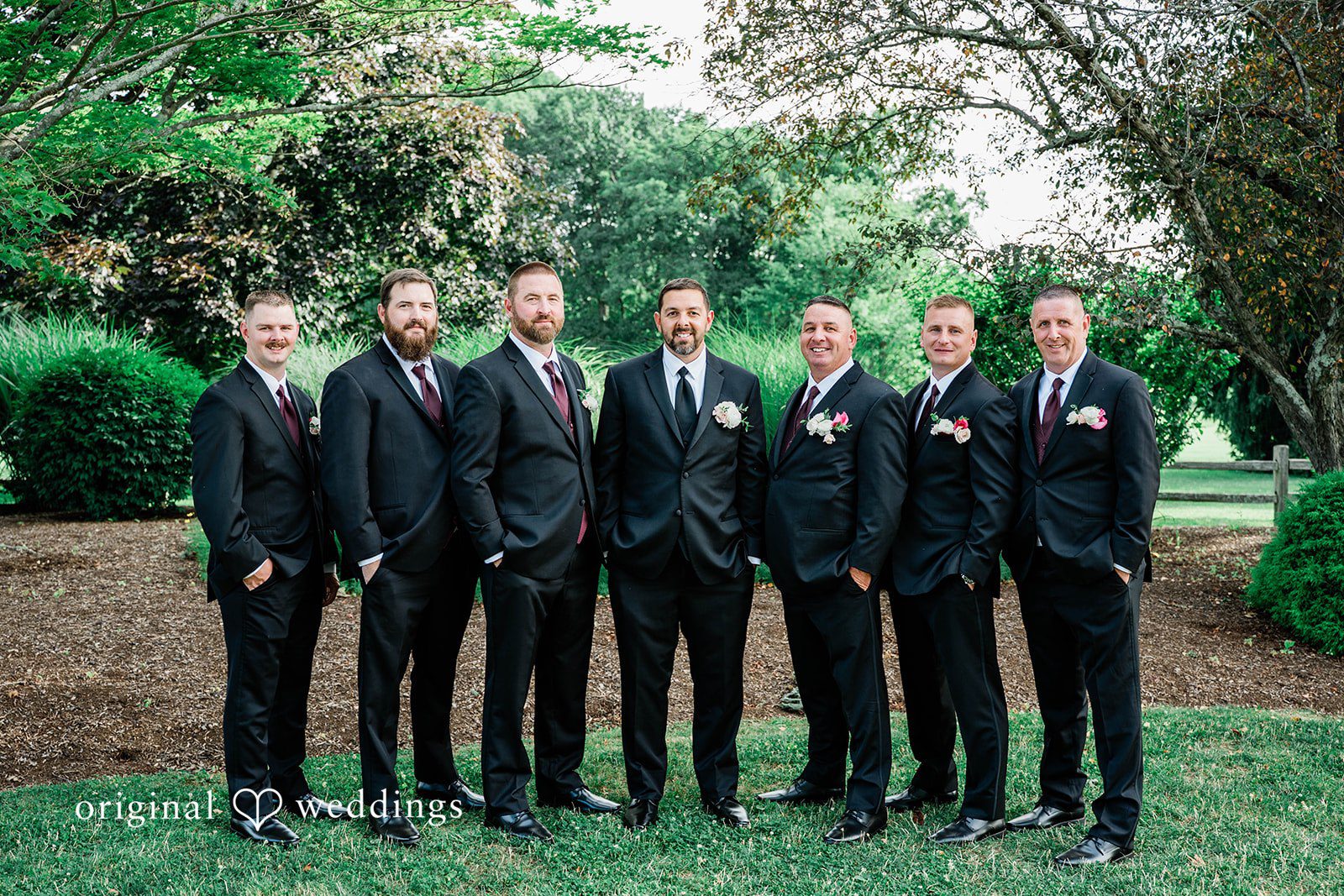 A group of groomsmen in black suits standing in a row on a lawn.