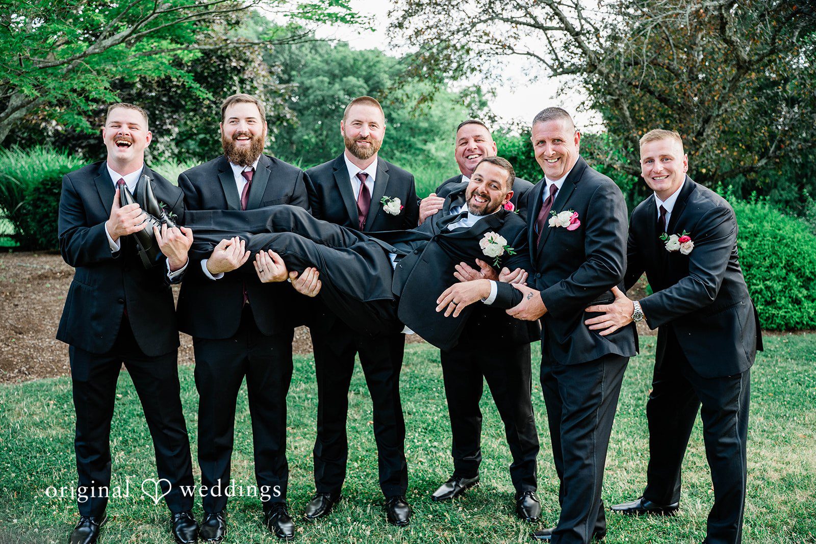 Groomsmen playfully lifting up the groom on a grassy field.