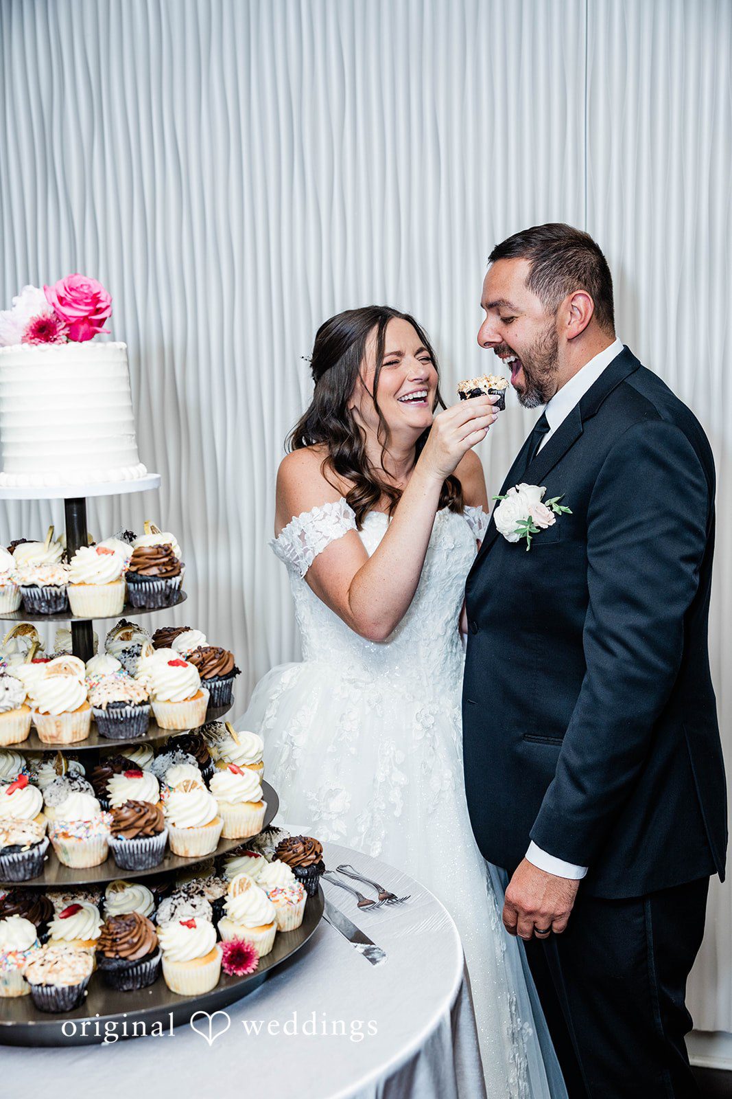A bride and groom feeding each other wedding cake next to a cupcake tower.