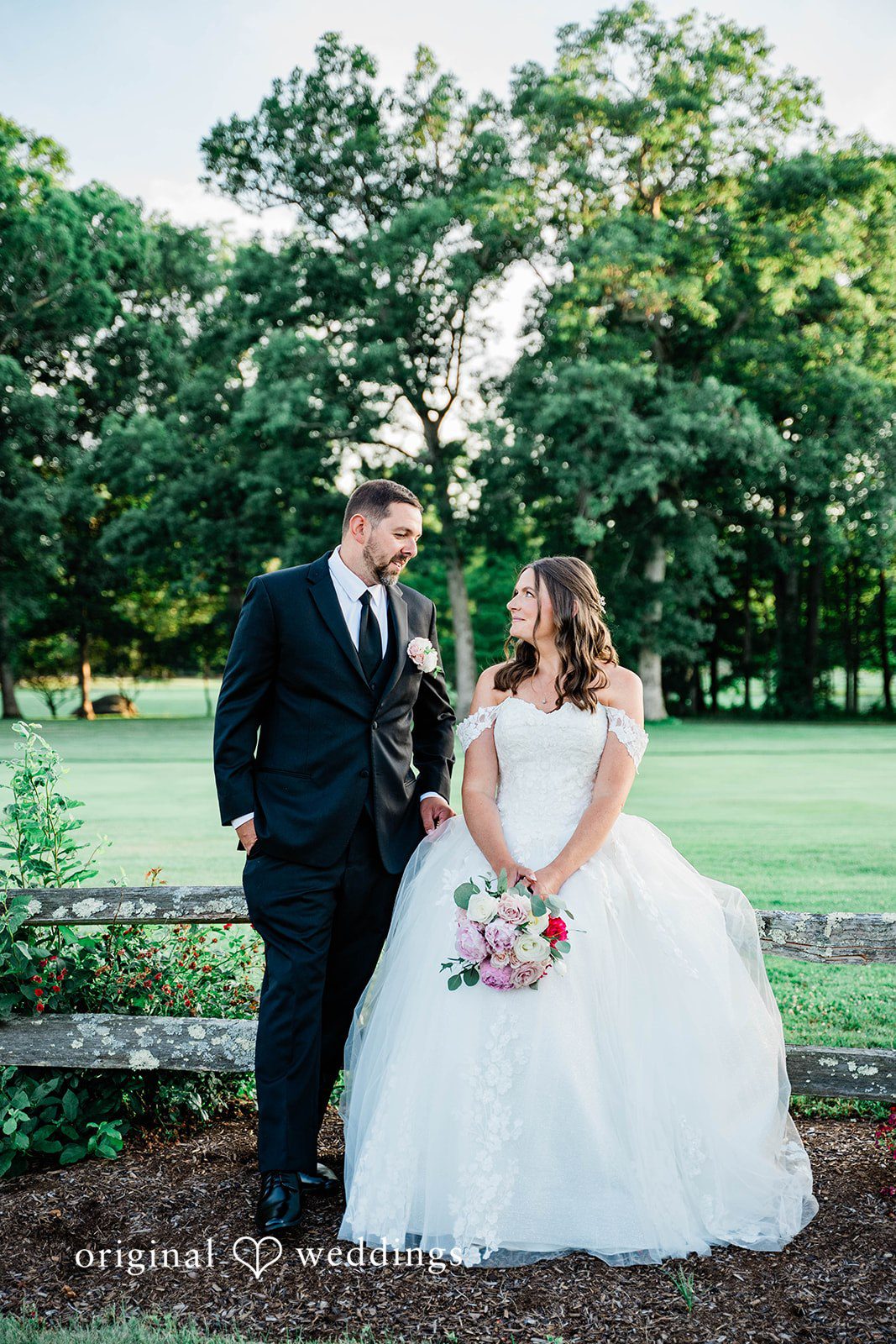 A bride and groom posing together on a green field at dusk.