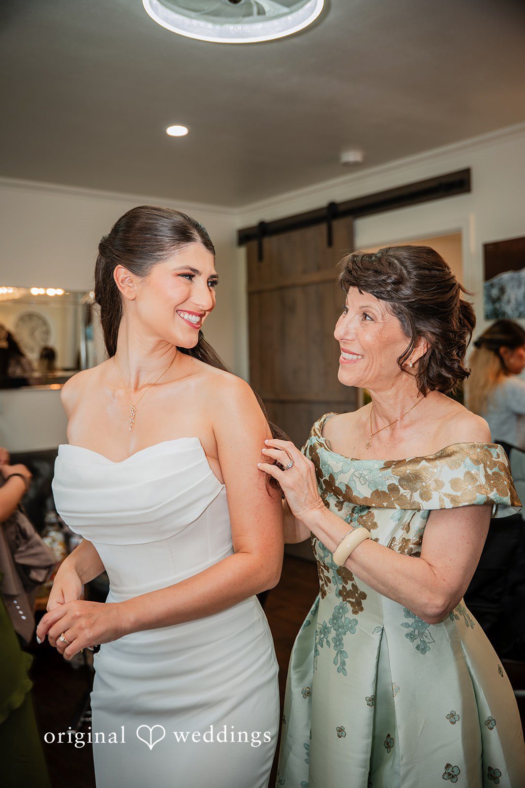 Zechariah + Tova A bride in a white wedding dress smiling at another woman dressed in a green patterned dress.