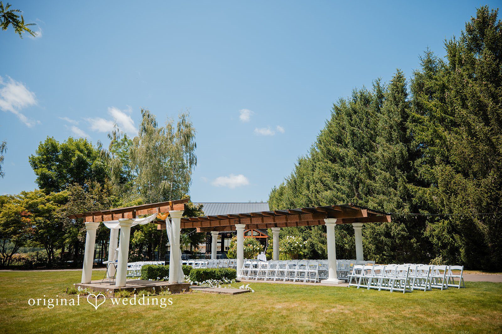 Zechariah + Tova A wide shot of a wedding ceremony venue with rows of white chairs facing a wooden pergola structure.