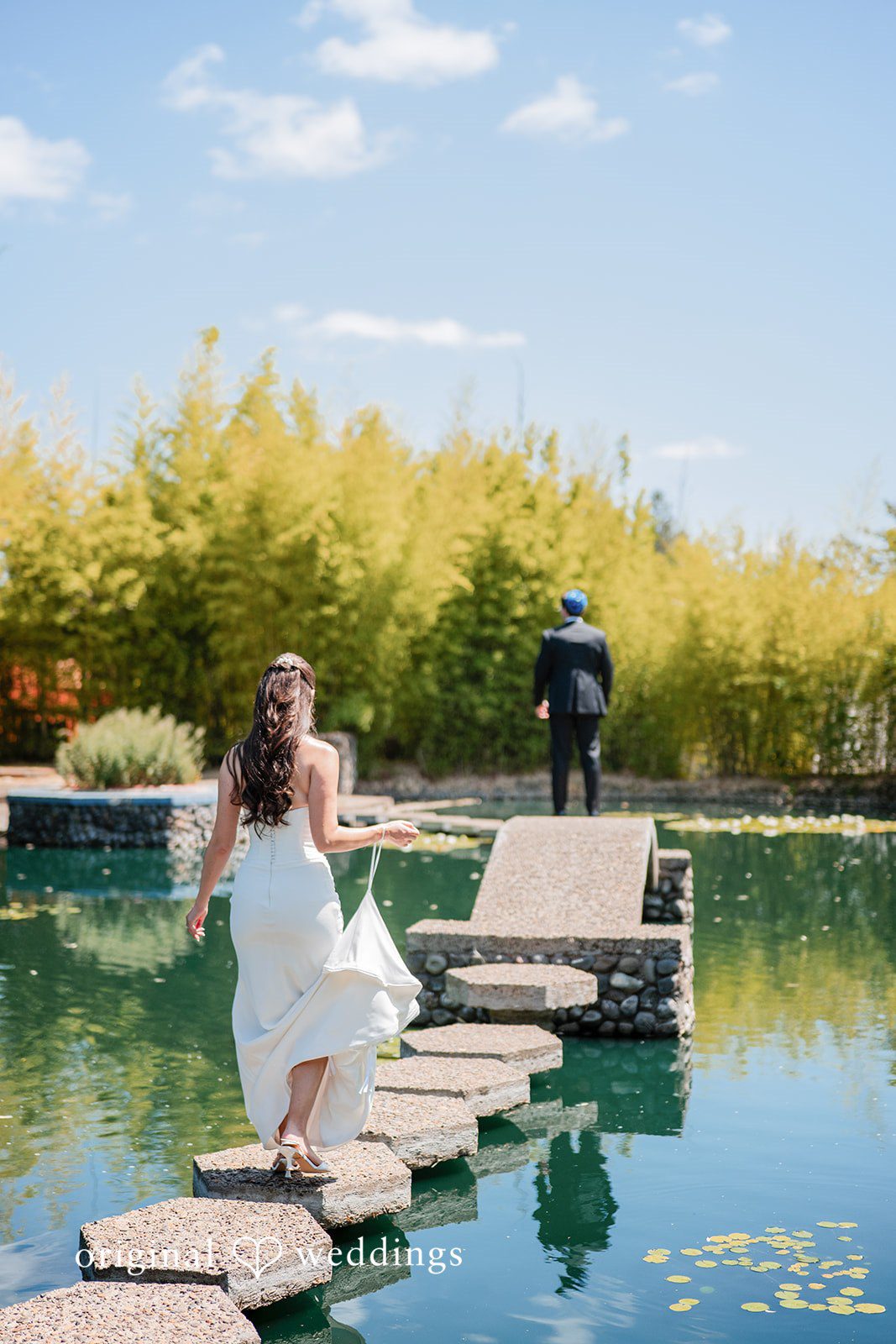 Zechariah + Tova A bride walking across stepping stones over a calm body of water toward a photographer.