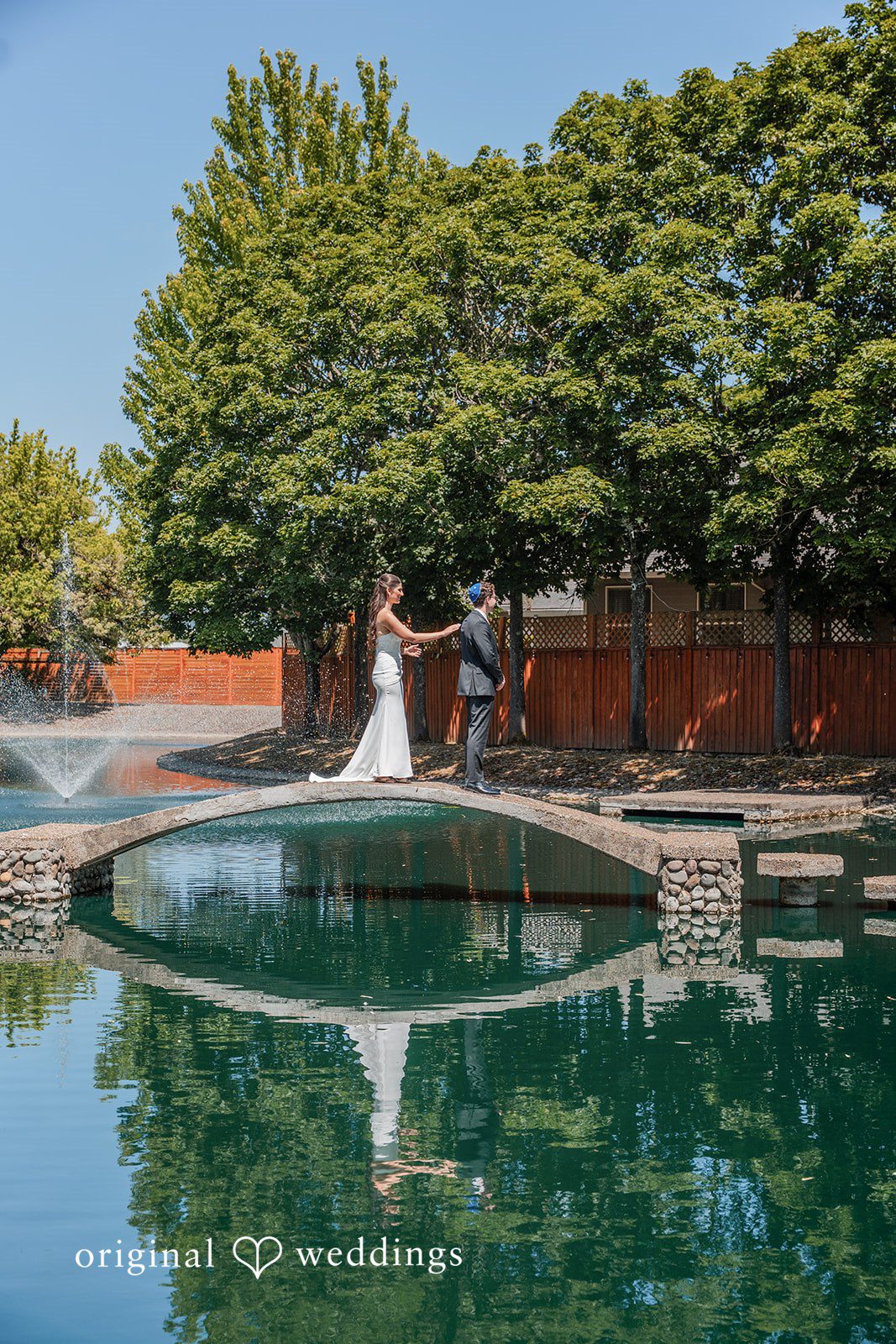 Zechariah + Tova A wide shot of a bride and groom standing on a stone bridge overlooking a large pond.