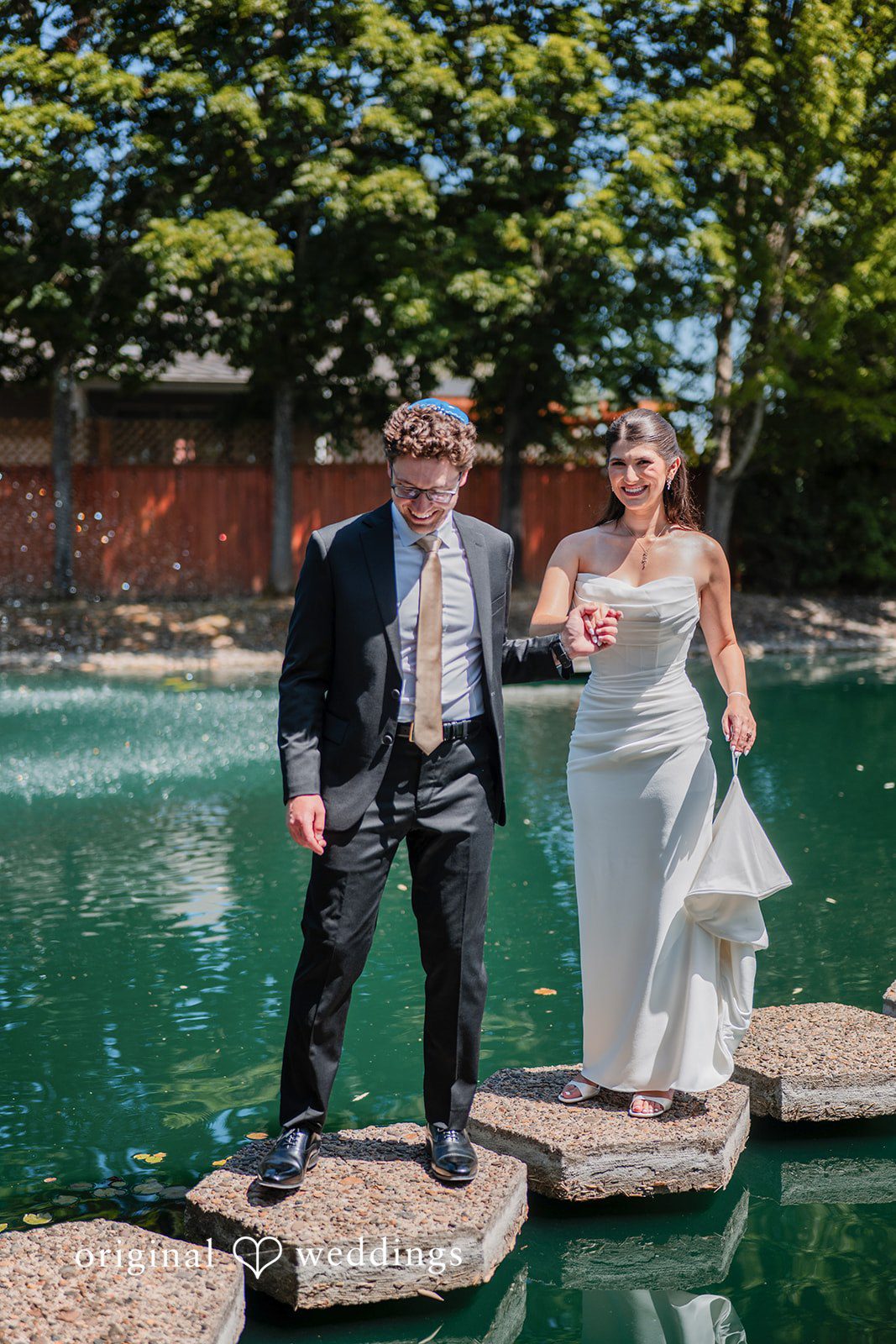 Zechariah + Tova A bride and groom holding hands while standing on a path of stepping stones across a pond.