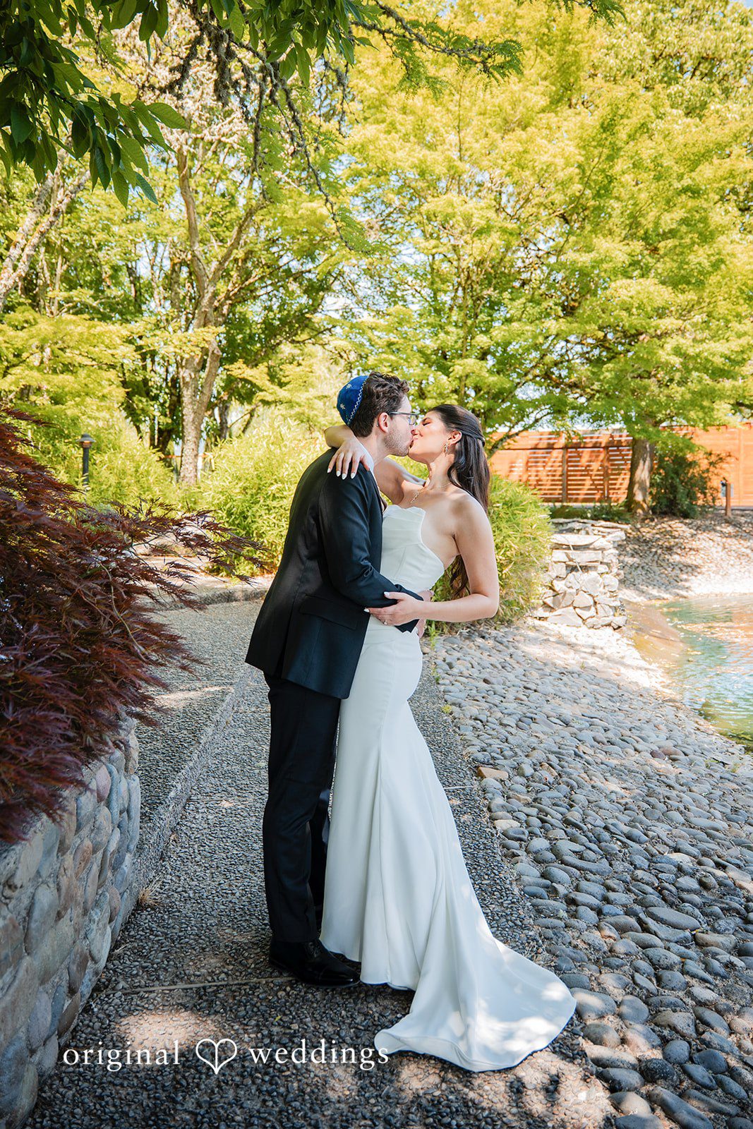 Zechariah + Tova A bride and groom walking hand-in-hand along a stone path with green trees in the background.