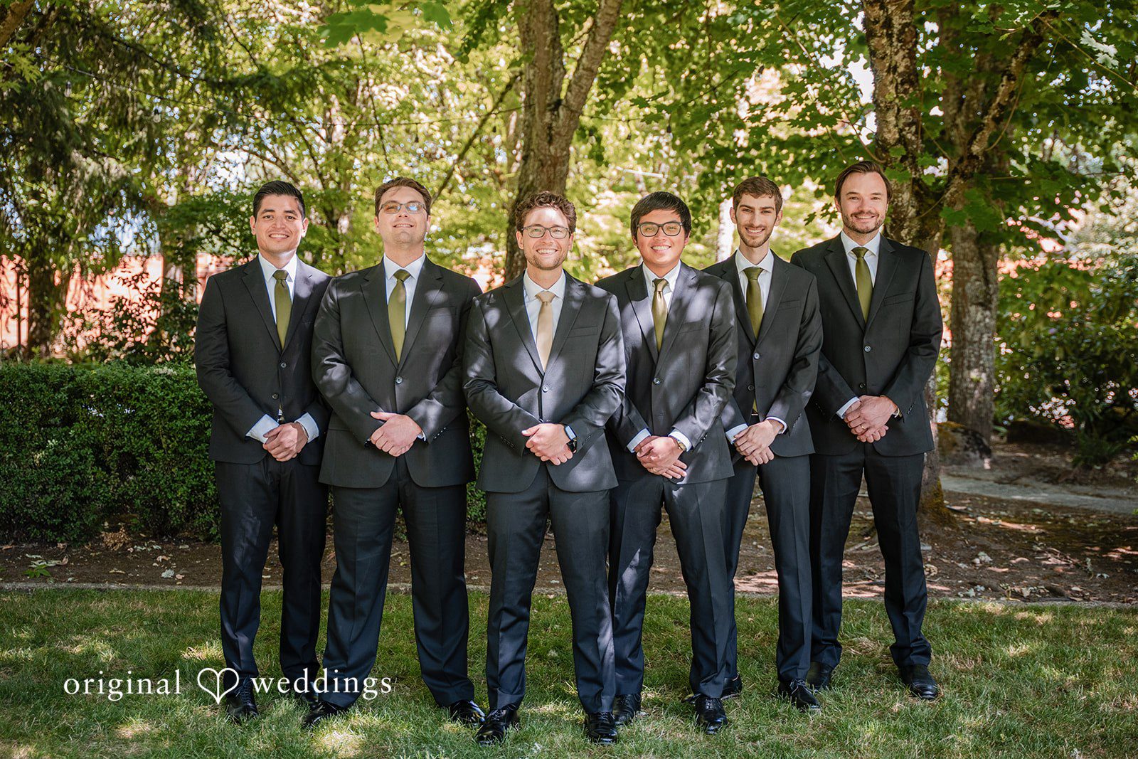 Zechariah + Tova Six men in matching dark suits posing together in a line in front of a wooded area.