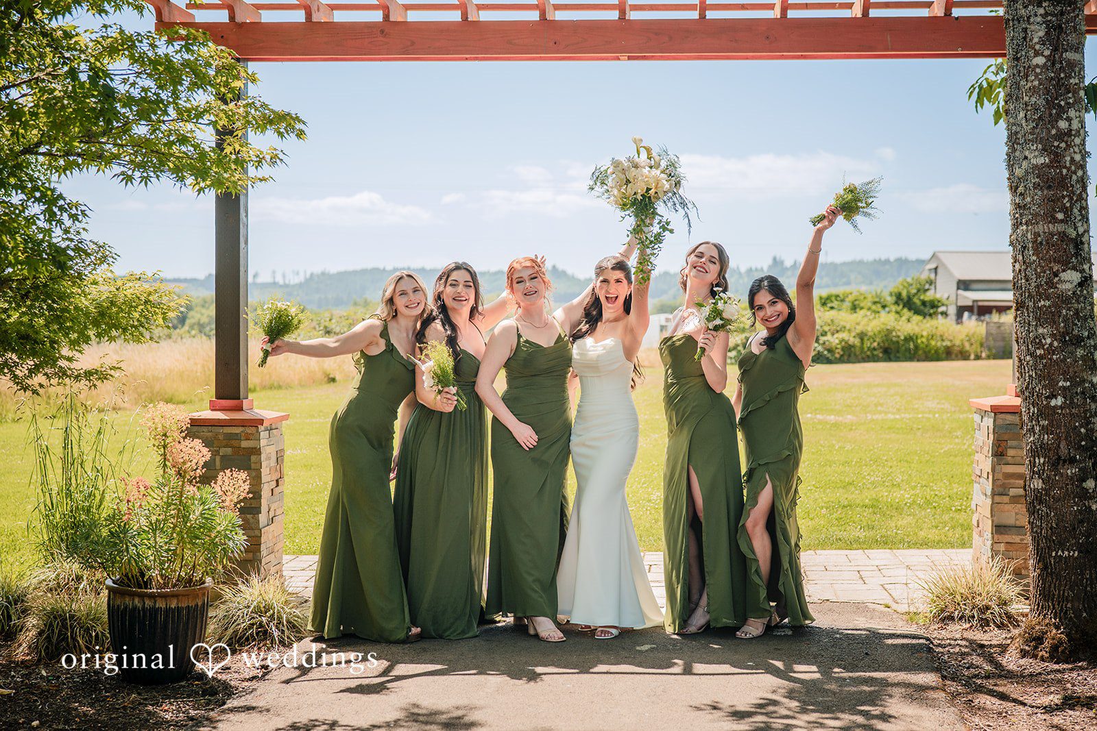 Zechariah + Tova A bride and five bridesmaids dressed in long green dresses posing together in front of a pergola.