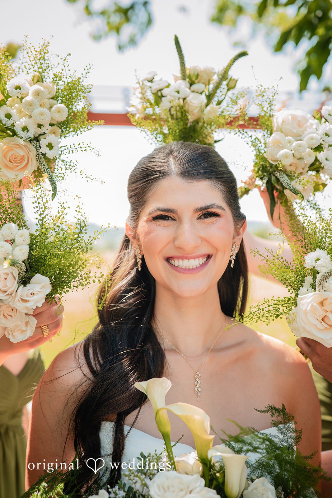 Zechariah + Tova A close-up portrait of a smiling bride holding a bouquet with white flowers and green foliage.