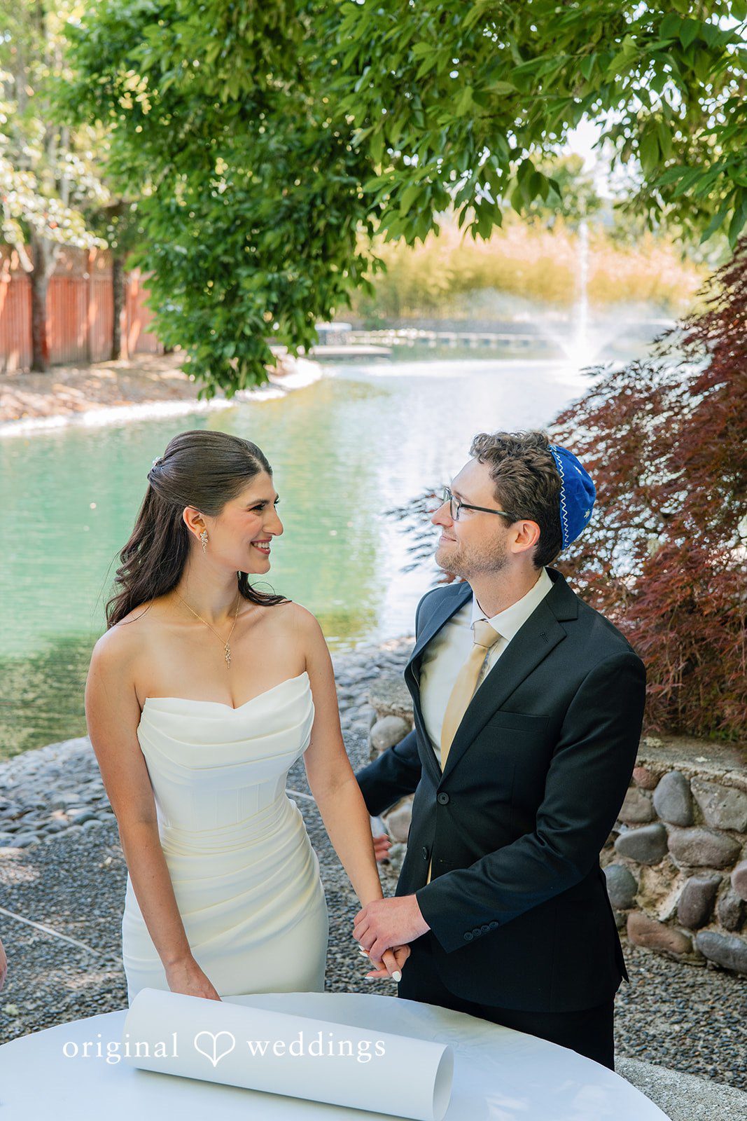 Zechariah + Tova A bride and groom standing together and smiling next to a pool of water.