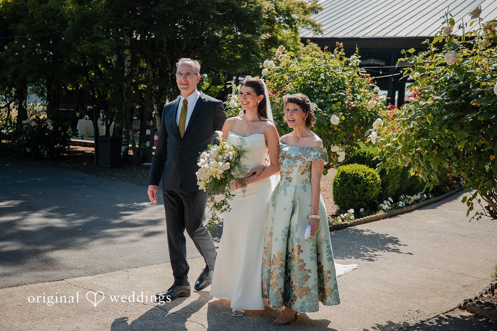 Zechariah + Tova A bride,man and women walking together down a paved path lined with trees and buildings.