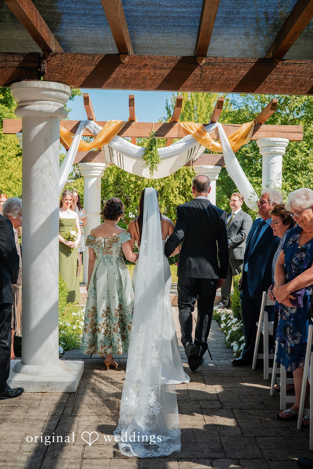Zechariah + Tova A bride seen from behind walking down a decorated wedding aisle towards guests under a pergola.