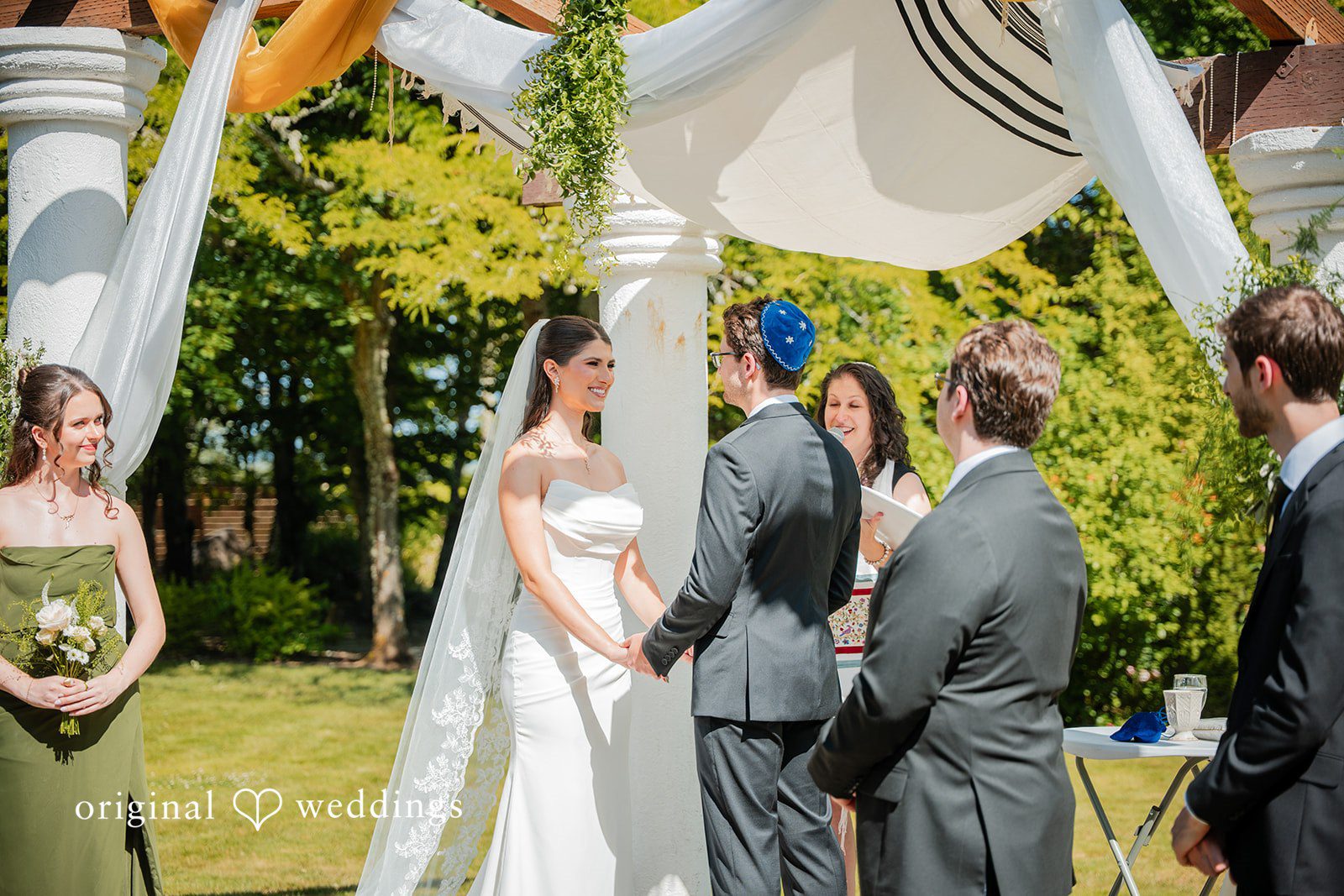 Zechariah + Tova A bride and groom during their wedding ceremony standing before an officiant under an umbrella.