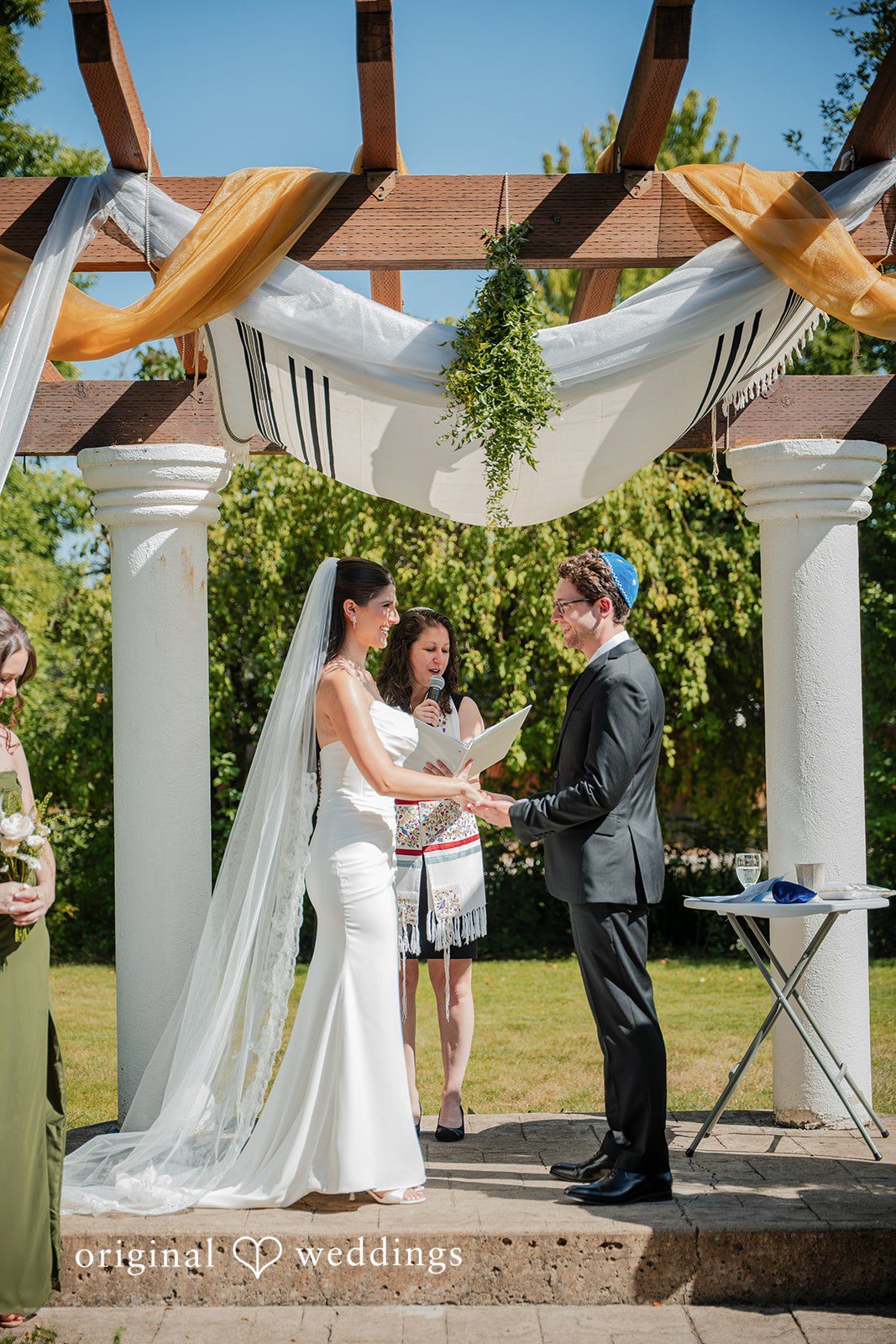 Zechariah + Tova A bride and groom holding hands during their wedding ceremony under a decorated white chuppah.