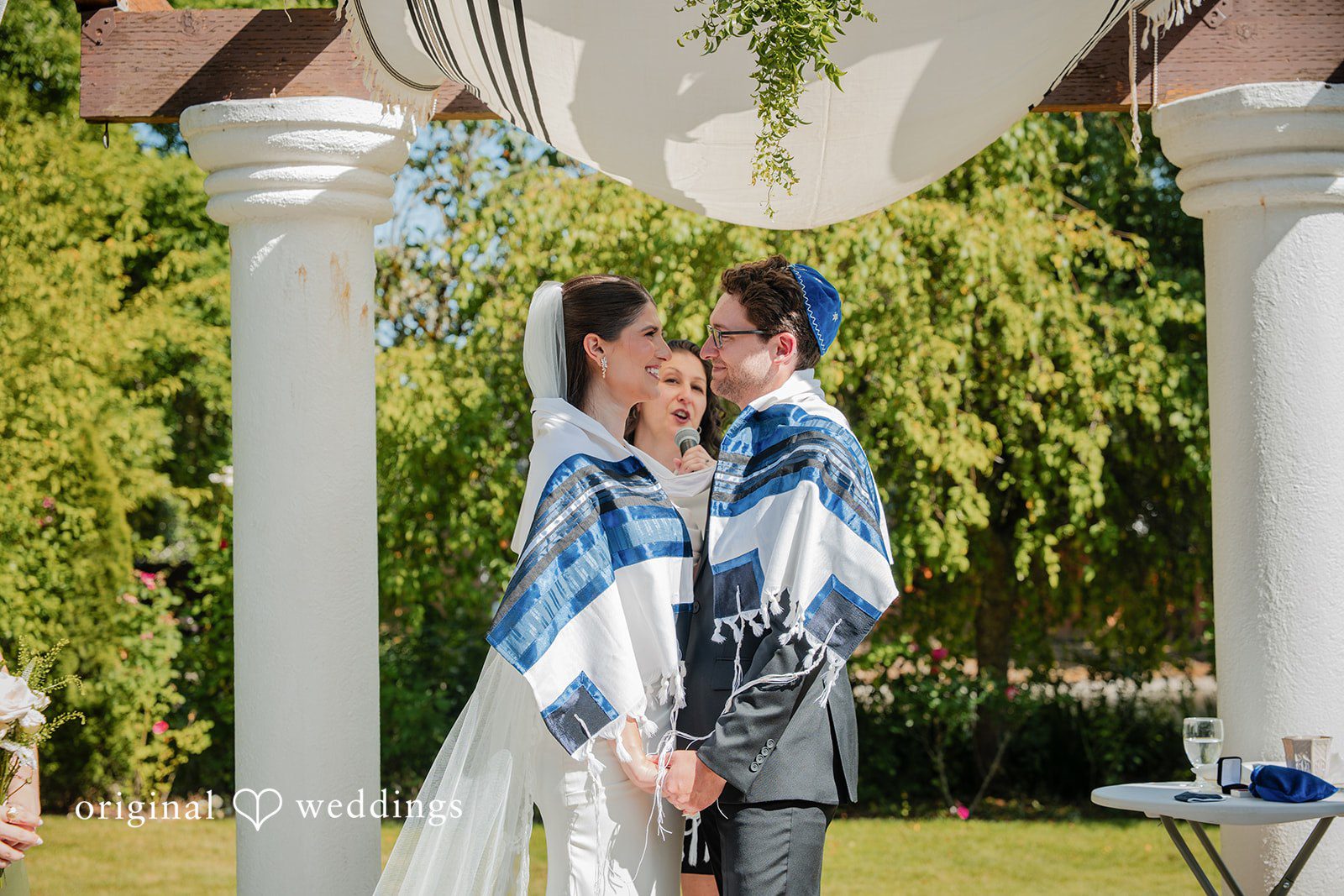 Zechariah + Tova A groom wearing a blue and white tallit standing next to his bride against green foliage.