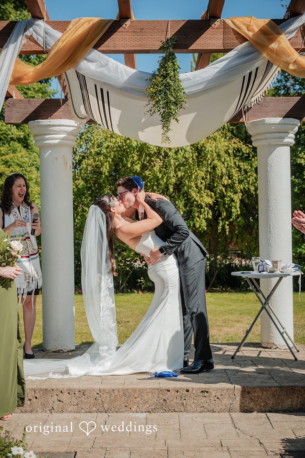 Zechariah + Tova A bride and groom sharing a kiss during their wedding ceremony under a decorated wooden canopy.
