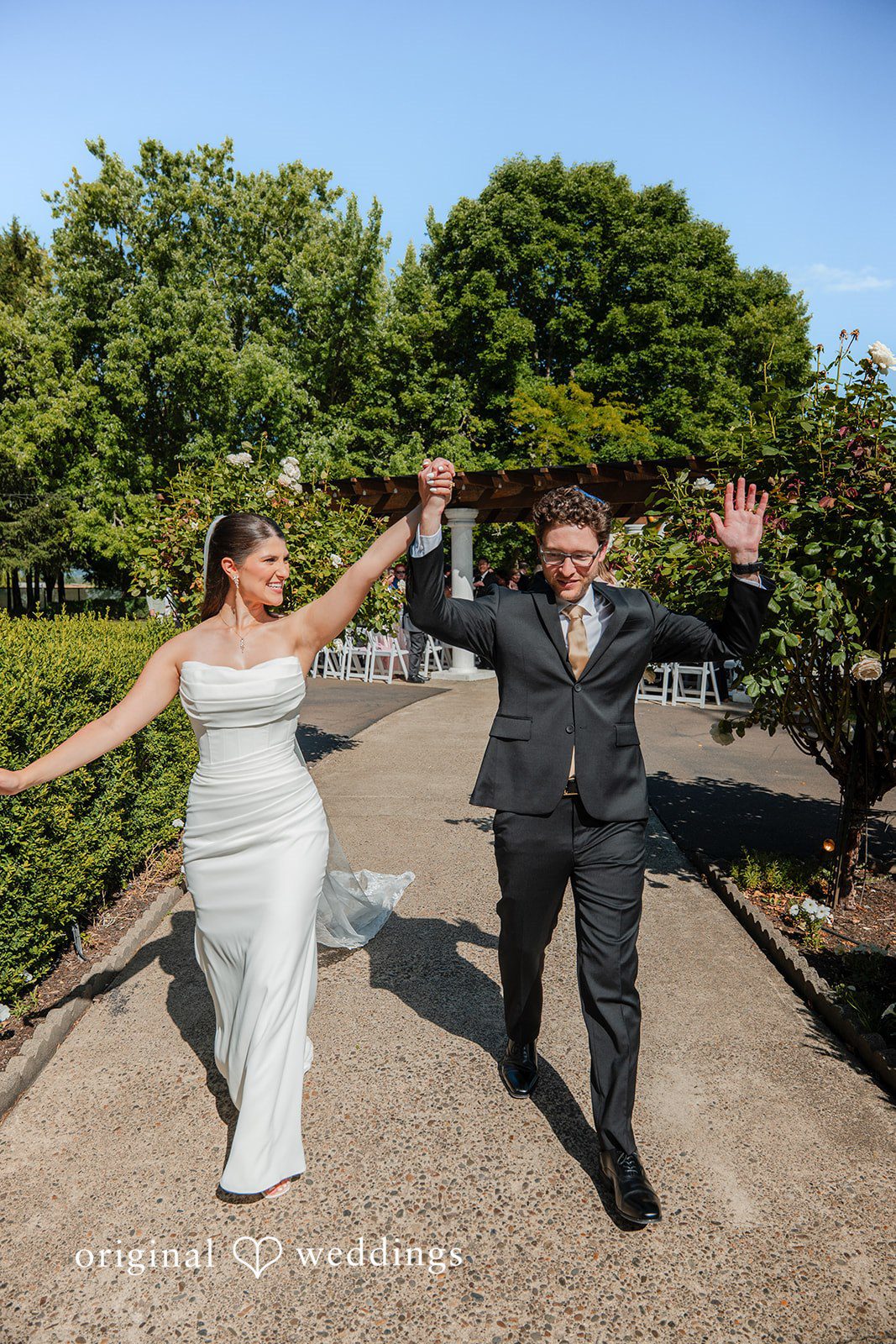 Zechariah + Tova A bride and groom holding hands and smiling while walking down a paved stone walkway.