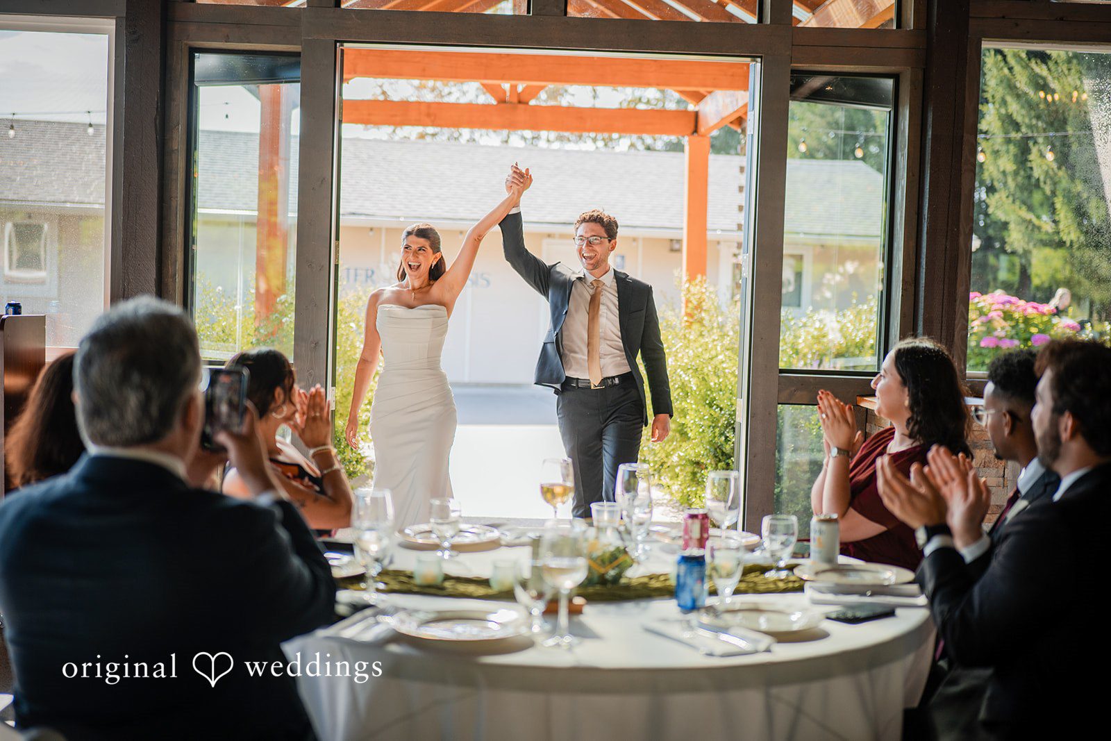 Zechariah + Tova A bride and groom raising their hands in celebration at their reception table surrounded by guests.