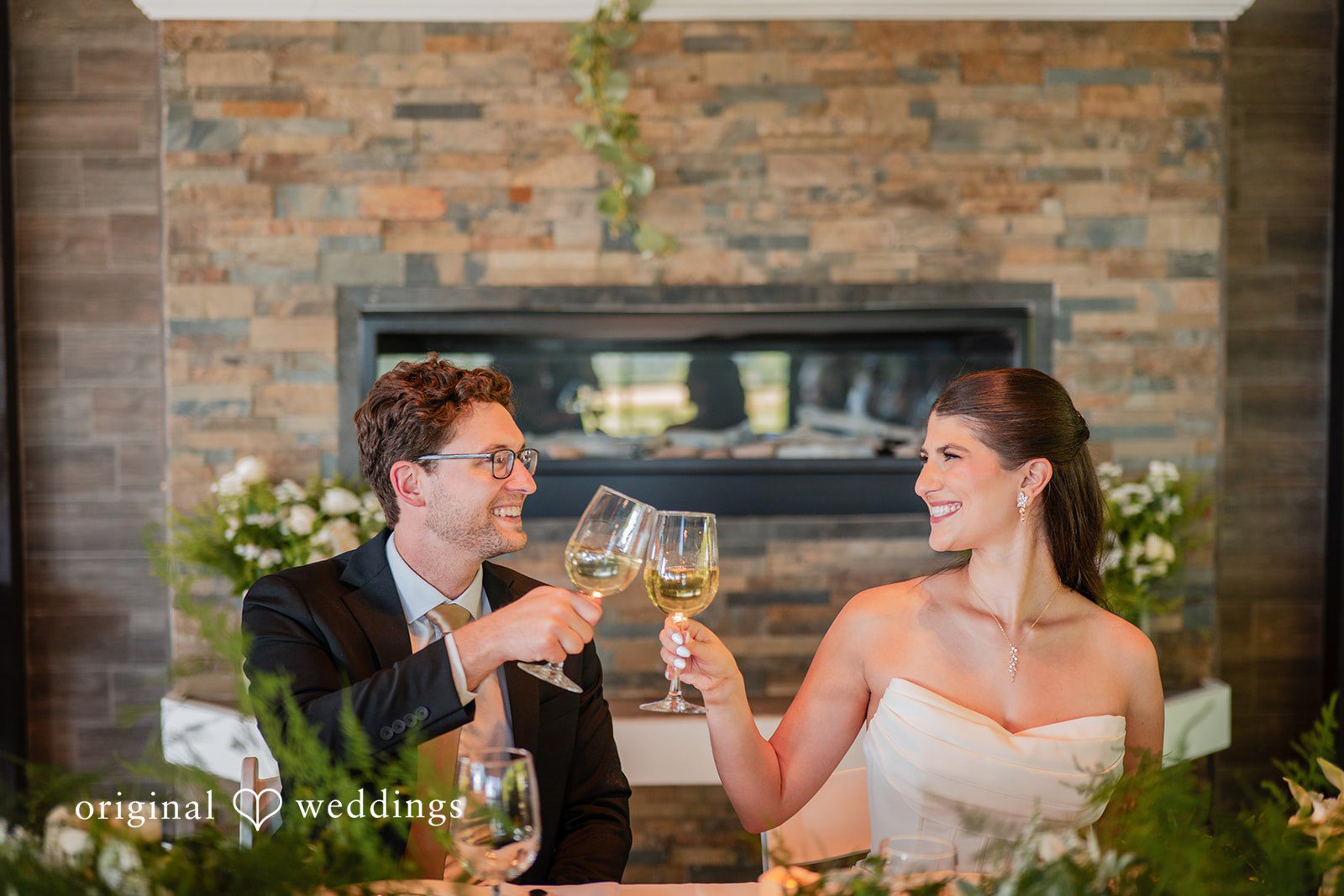 Zechariah + Tova A bride and groom sitting together and raising a toast with wine glasses in front of a fireplace.