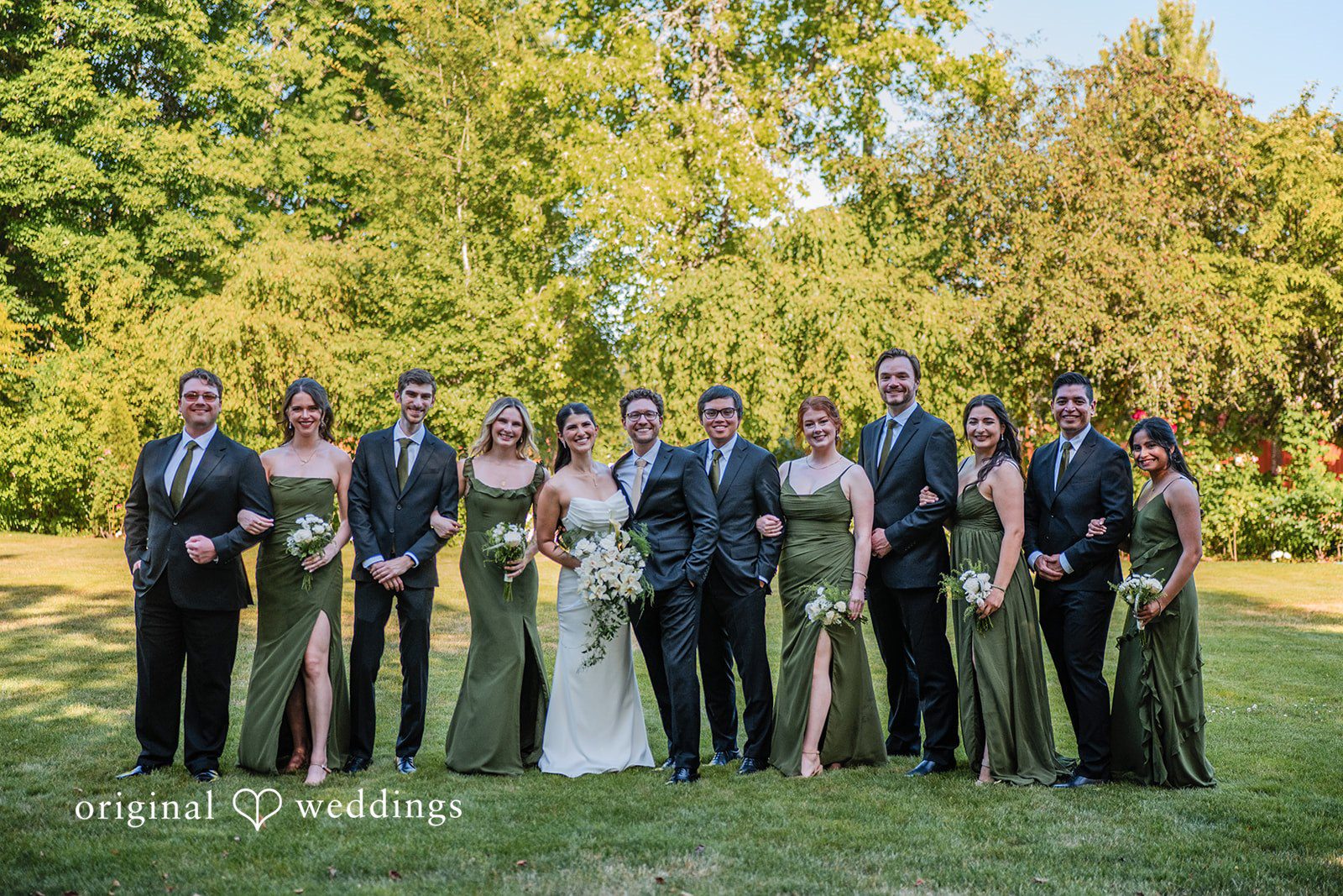 Zechariah + Tova A large wedding party consisting of bridesmaids and groomsmen posing together in a line on a lawn.