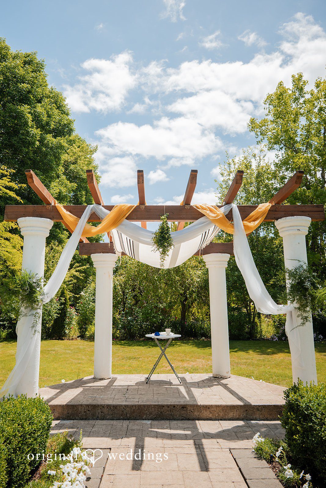 Zechariah + Tova A wedding ceremony altar with white pillars and hanging fabric drapes under a bright sky.