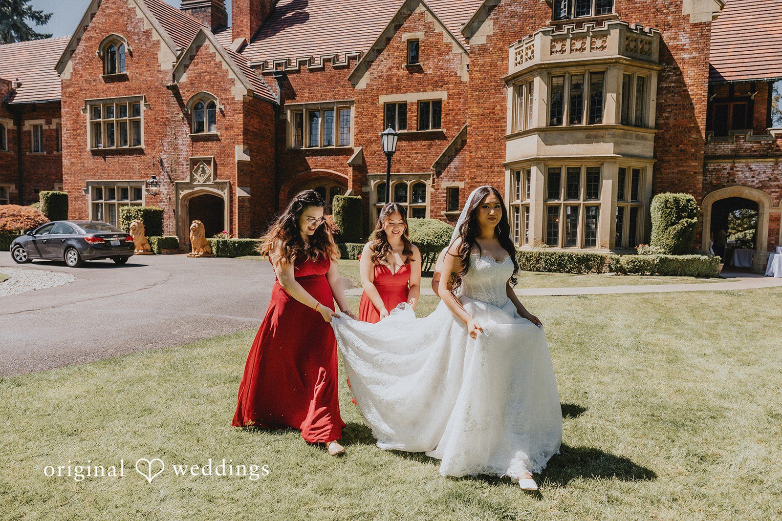Sharmaine + Ronald Two bridesmaids in red dresses hold up the train of a bride's gown as they walk across a lawn in front of a brick estate.