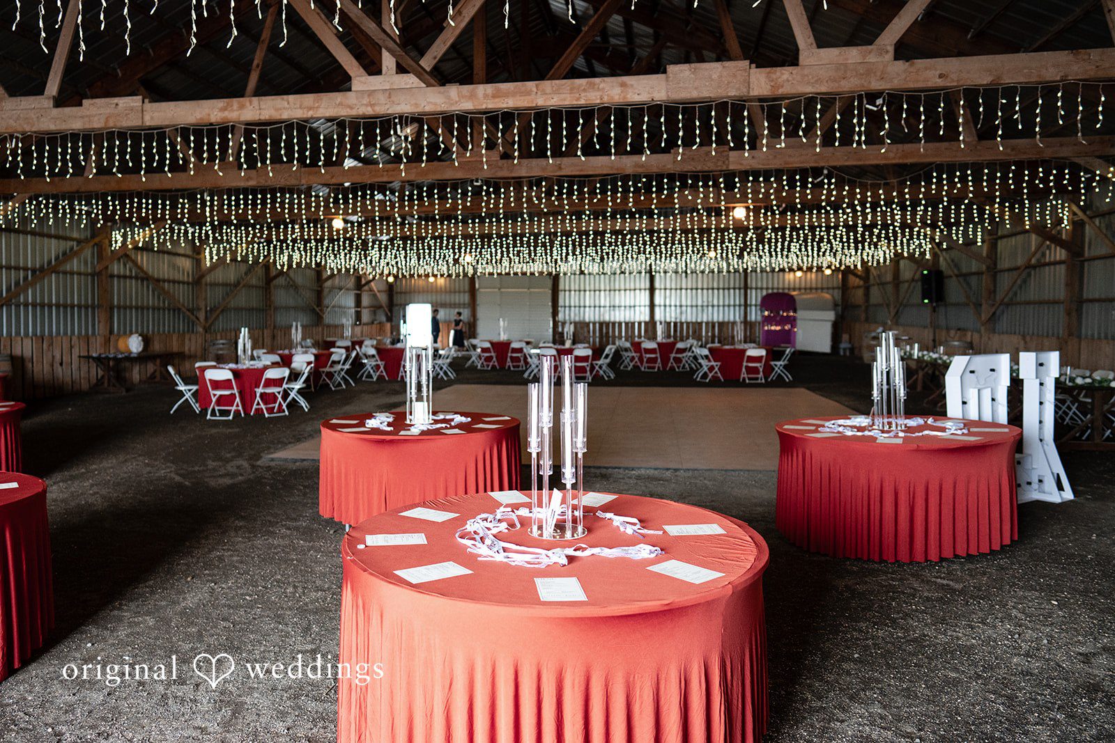 Kayla + Dillon A wide view of a rustic reception venue arranged with round tables draped in red tablecloths.