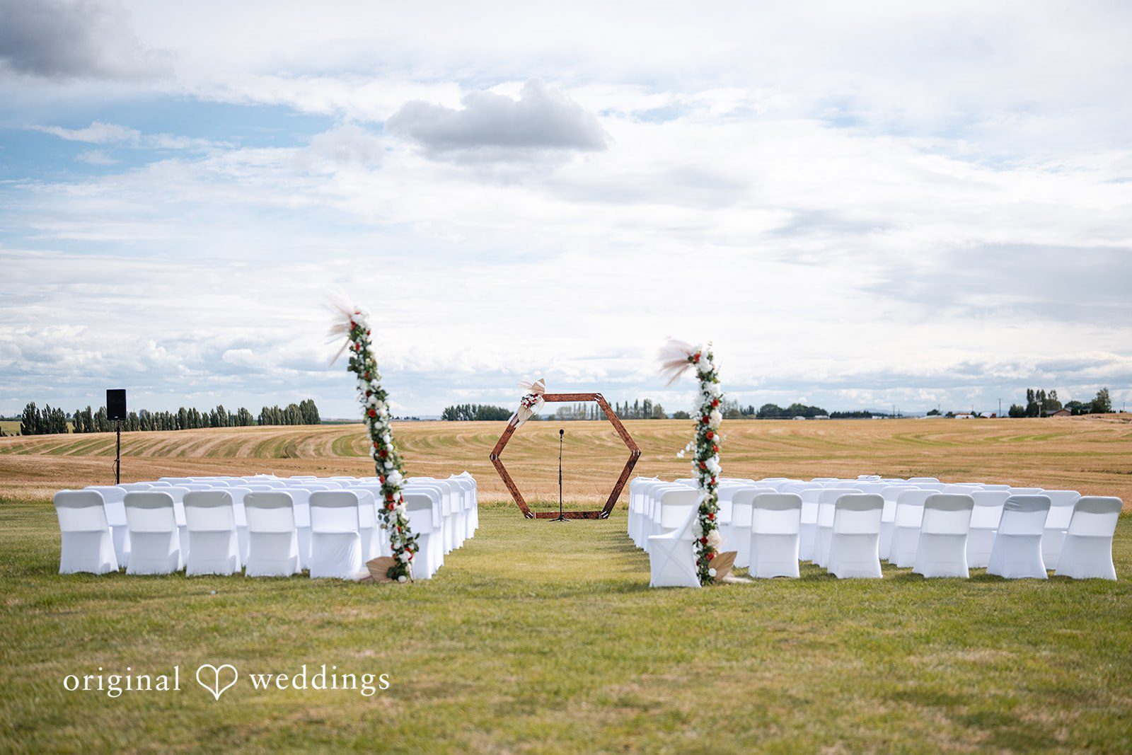 Kayla + Dillon A hexagonal wooden wedding arch set up in an open grassy field with white chairs.