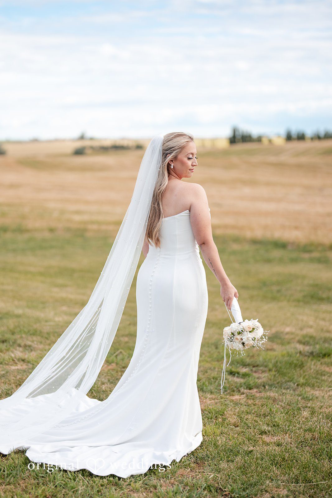 Kayla + Dillon A bride in a long flowing veil walking across a wide open field.