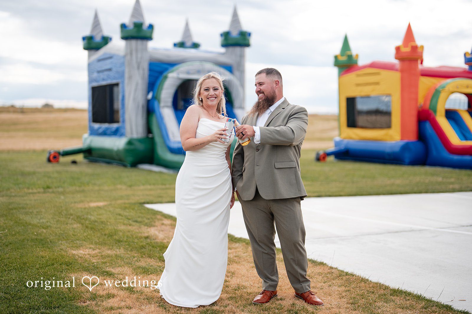 Kayla + Dillon A bride and groom posing together on a concrete path with a colorful inflatable bounce house behind them.