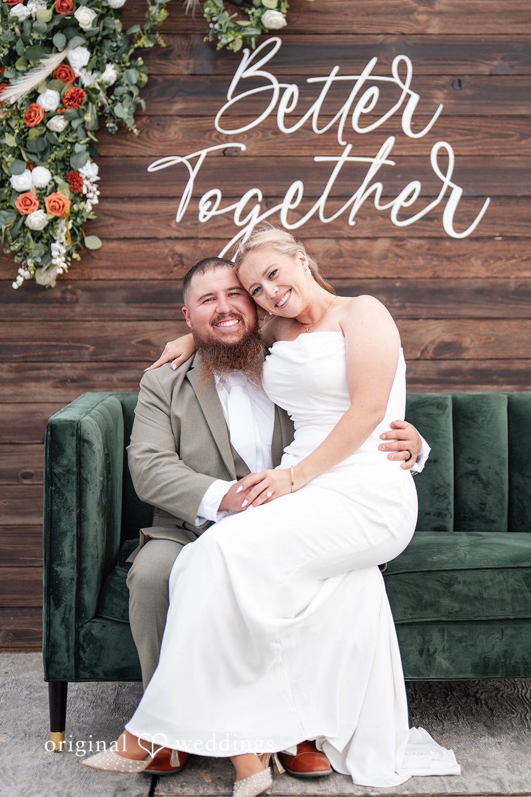 Kayla + Dillon A bride sitting on her groom's lap on a green velvet couch in front of a rustic wooden backdrop.