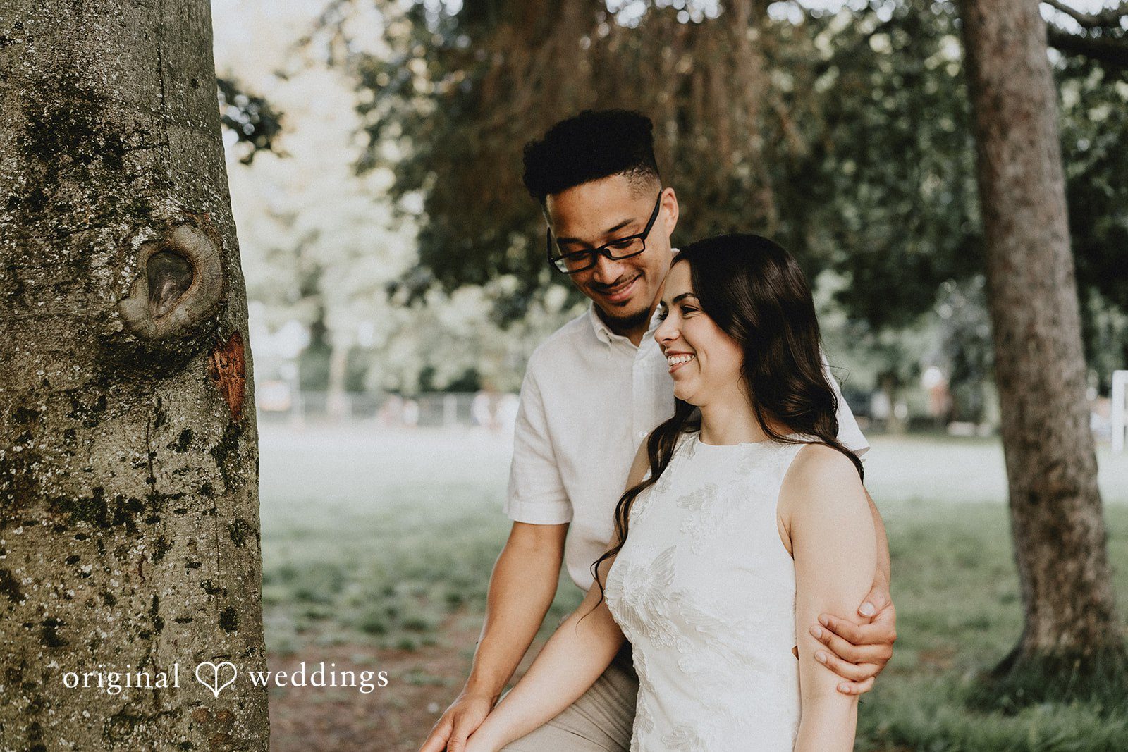 Man standing behind a woman while holding her waist among green trees.