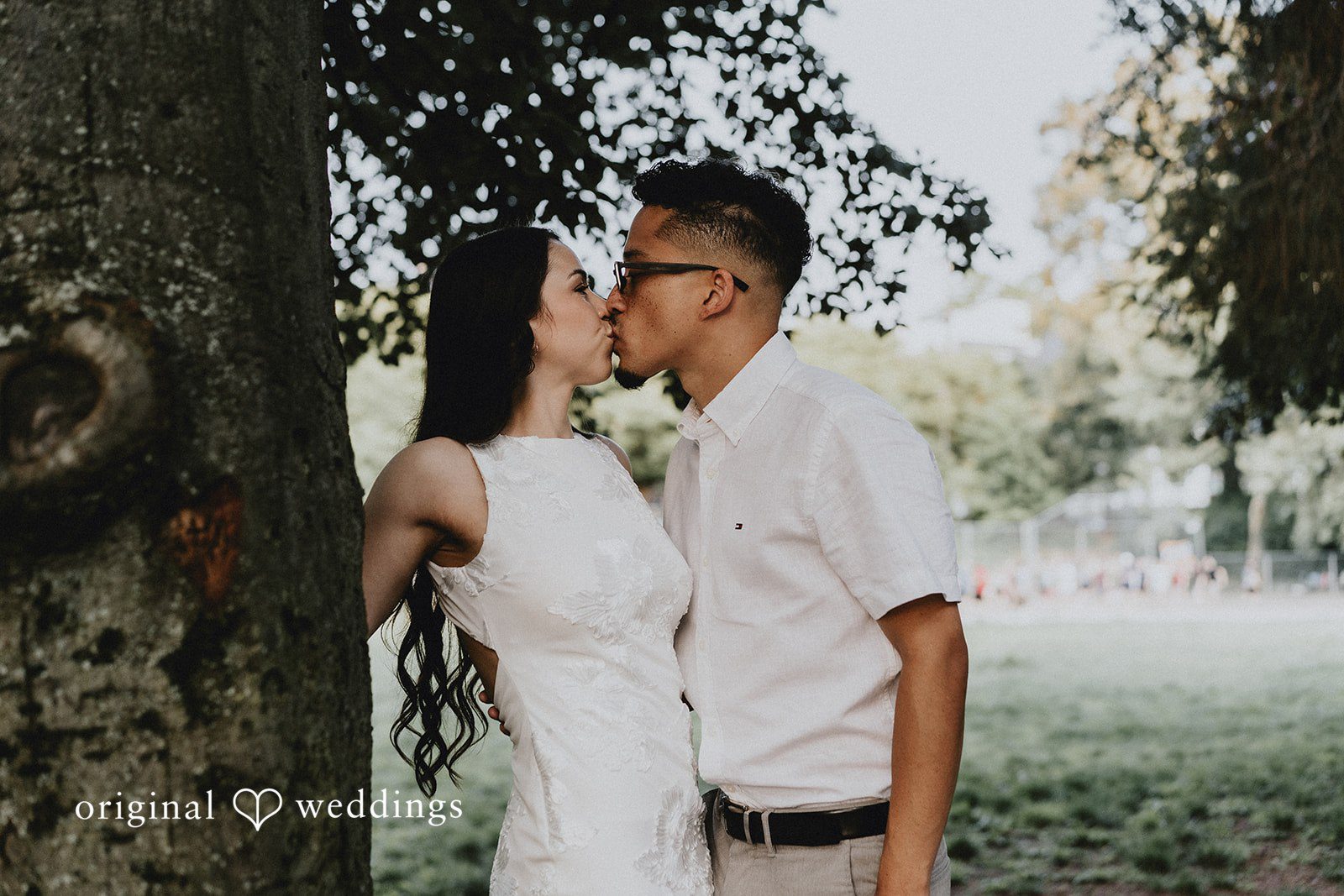 Couple sharing a romantic kiss while standing under a large tree canopy.